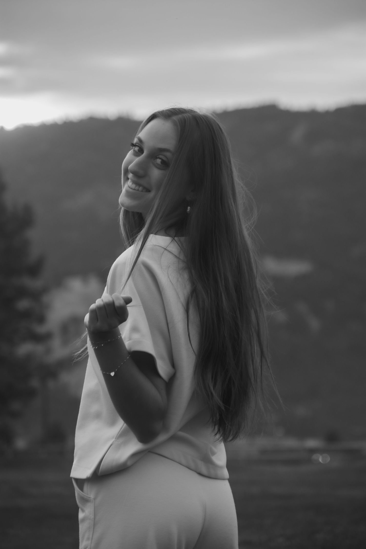 Black and white portrait of a smiling young woman with long hair posing outdoors with mountains in the background.