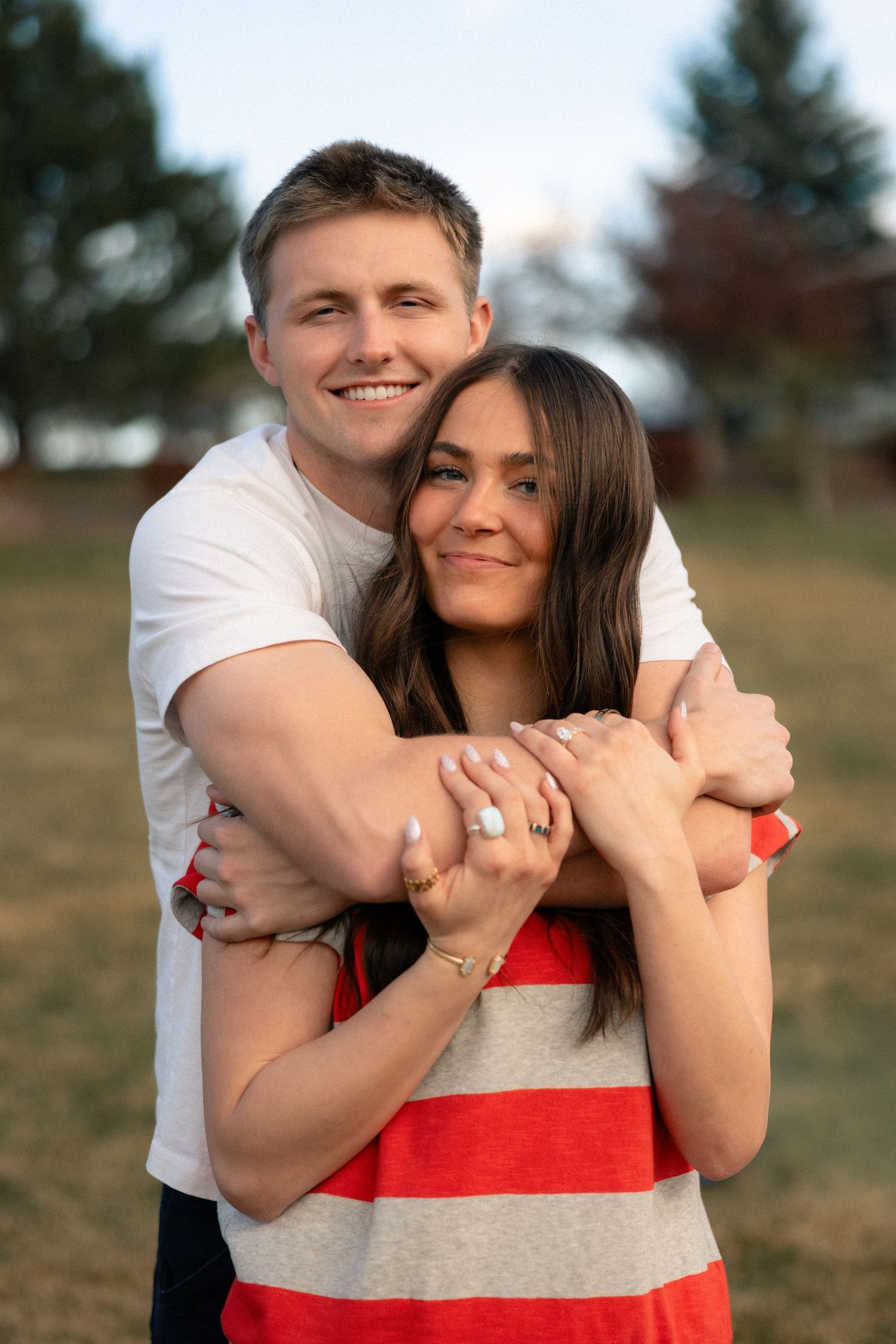 A smiling couple embraces outdoors, man in white tee hugging woman in red striped top, showcasing an engagement ring.