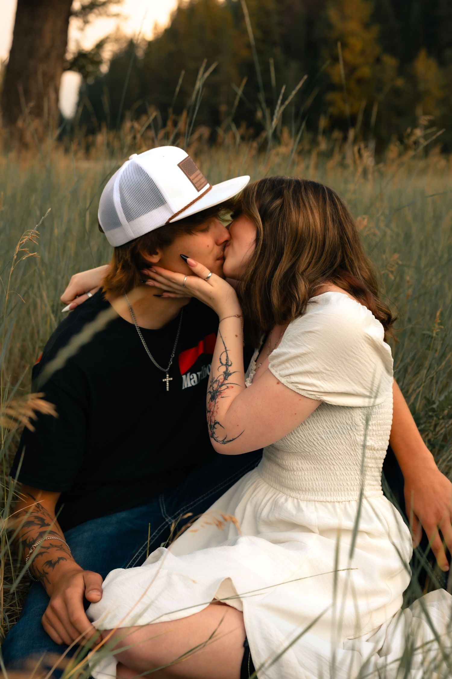 A couple shares a romantic kiss while sitting in a tall grass field, woman in white dress and man in black shirt and cap.