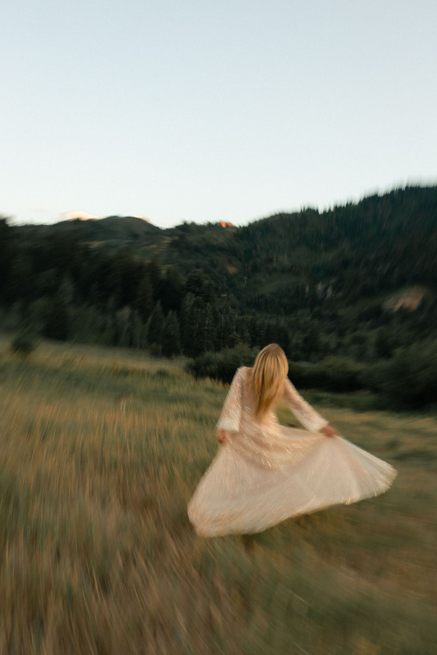 A woman in a flowing white dress runs through a golden meadow with lush green mountains in the background.