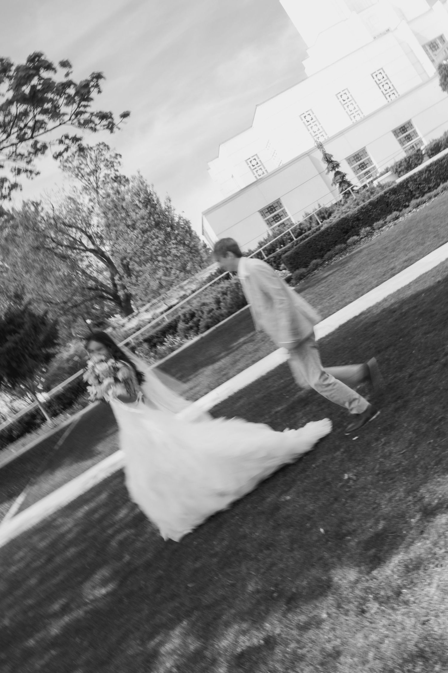 Black and white photo of a bride and groom running across a lawn outside a white building on their wedding day.
