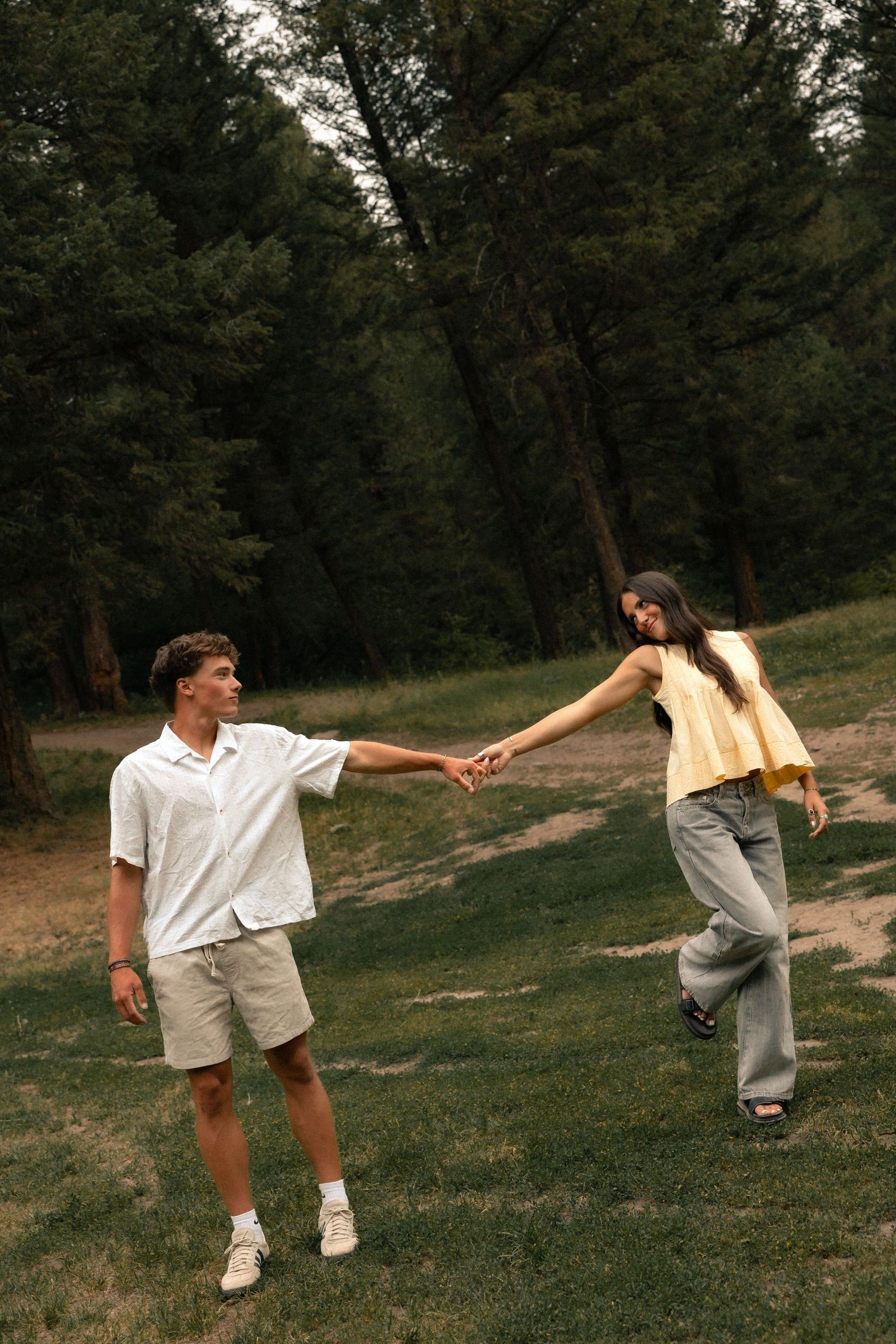 A young couple holds hands in a lush green forest park, the man in white shirt and the woman in a yellow top.