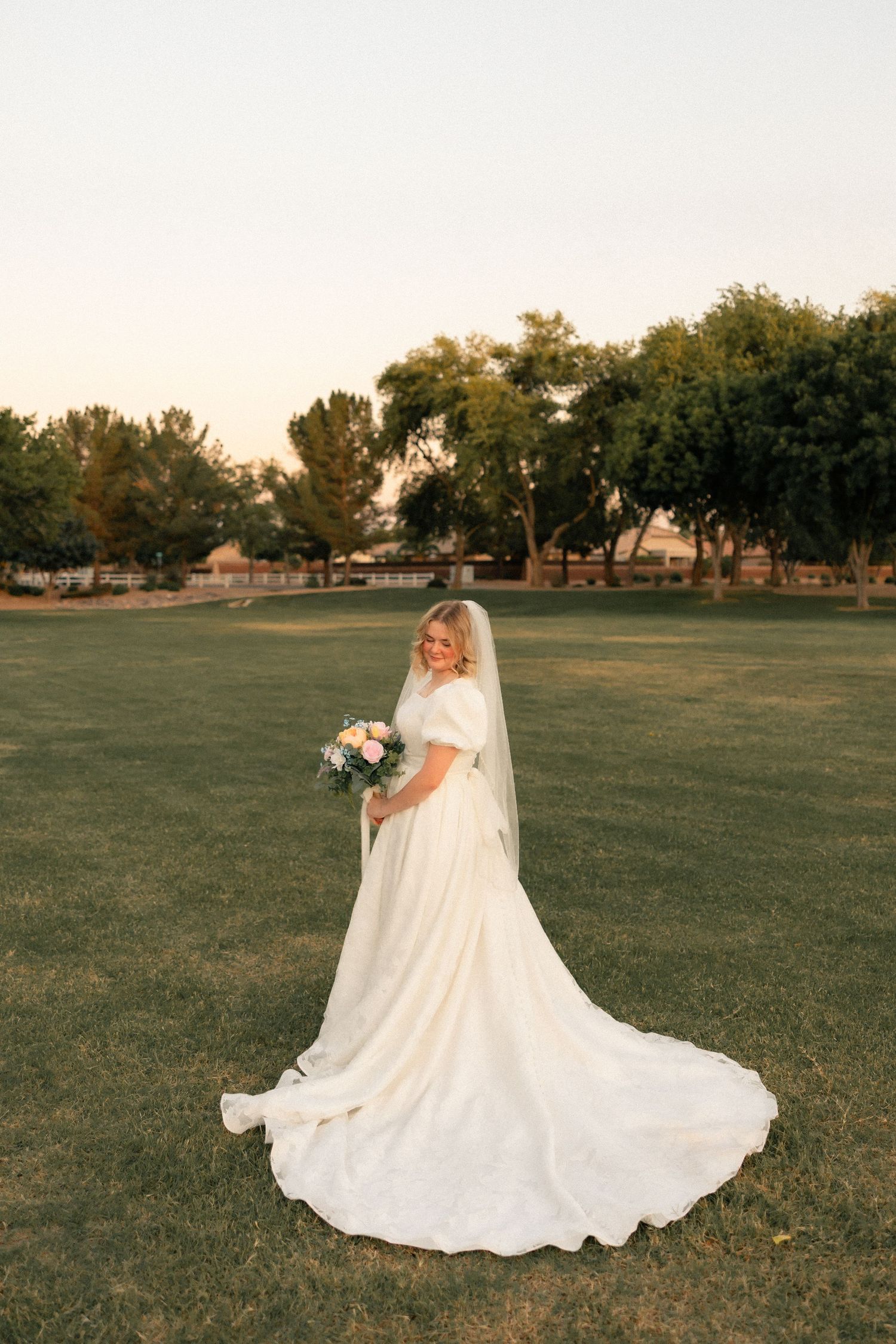 Bride in white gown with long train holds bouquet on green lawn at sunset with trees in background.