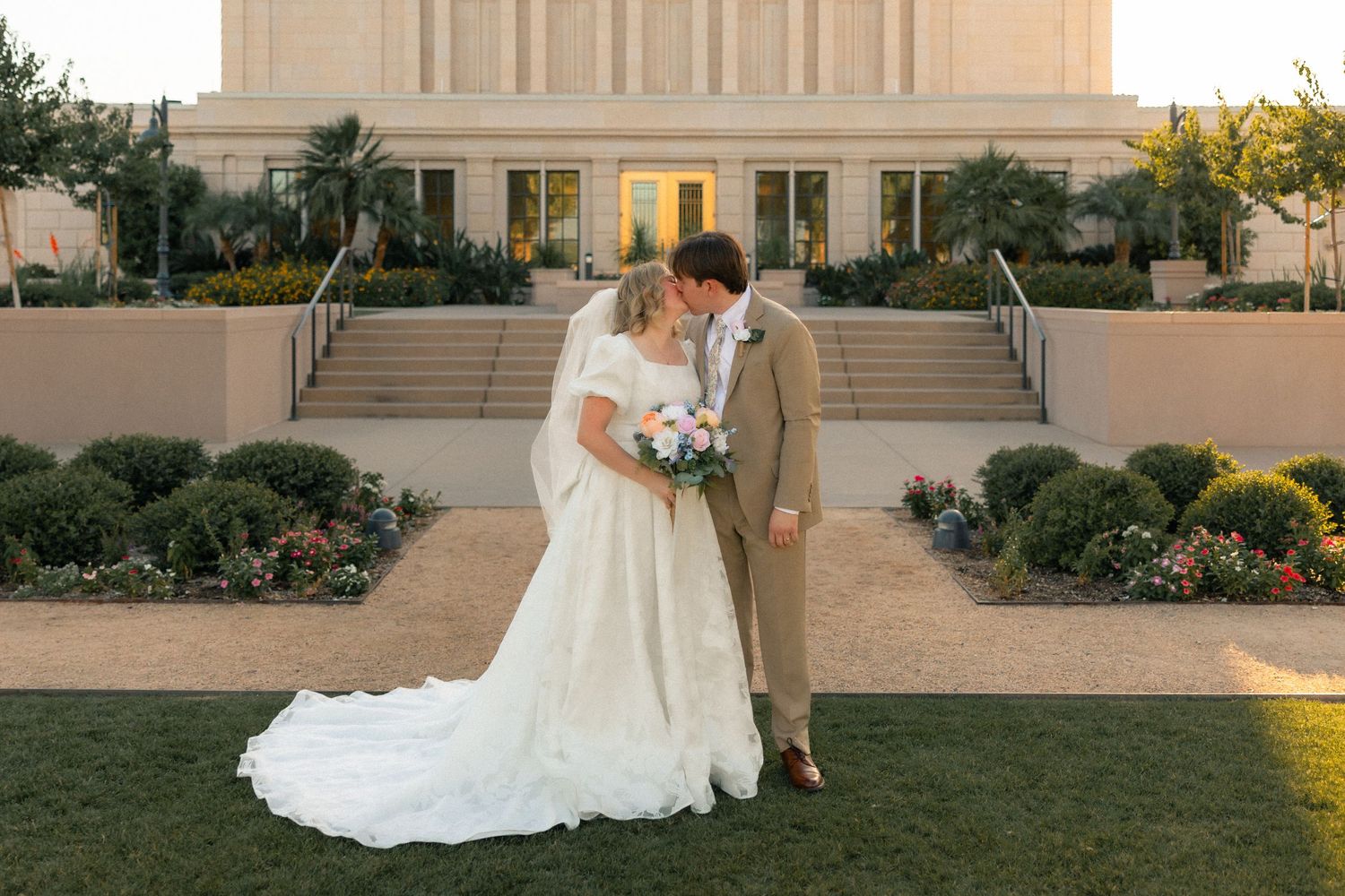 Bride and groom share a kiss in front of a grand temple, bride in elegant white gown holding a bouquet.