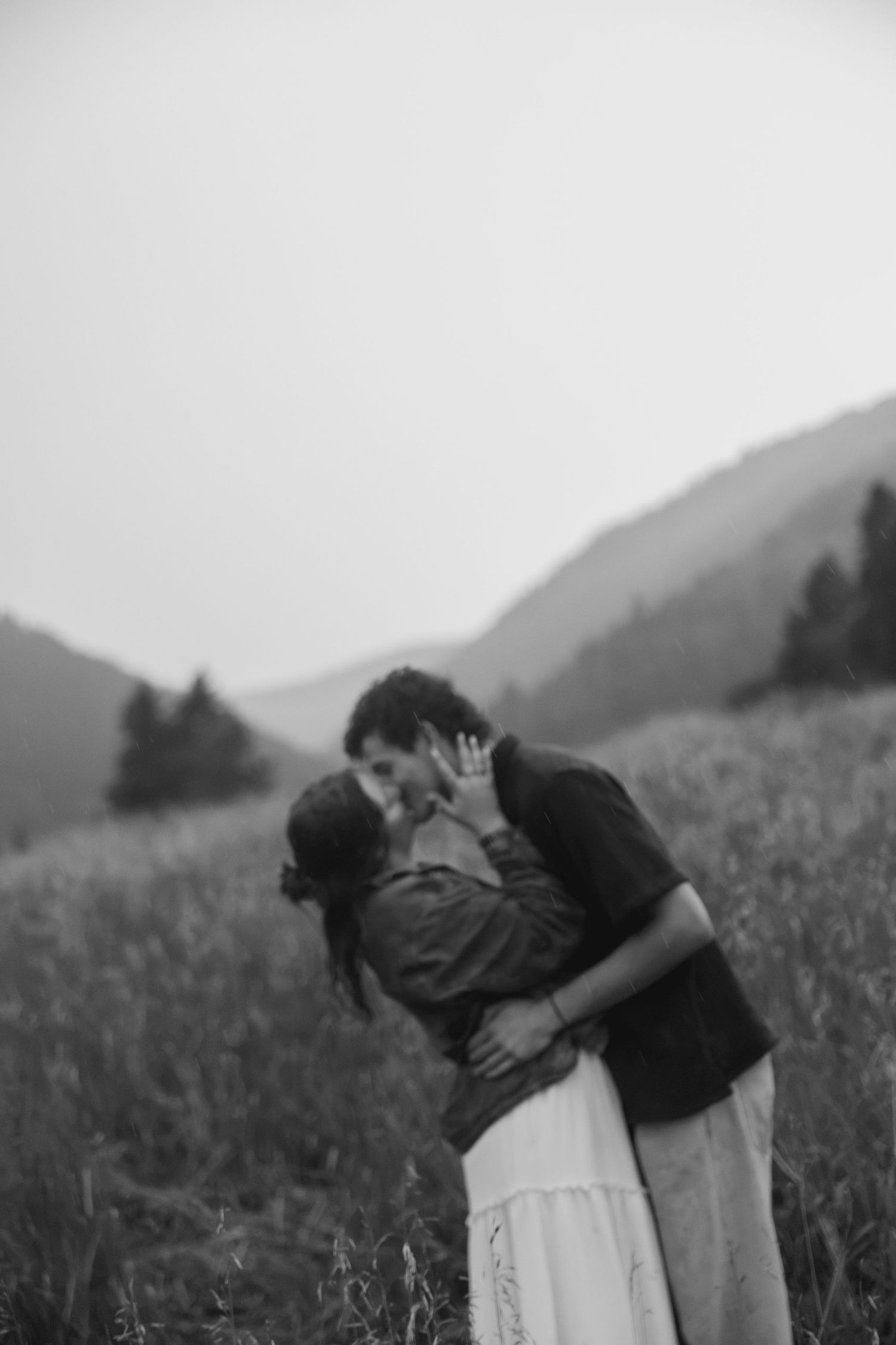 Black and white photo of couple sharing romantic kiss in misty mountain field, woman in white skirt.