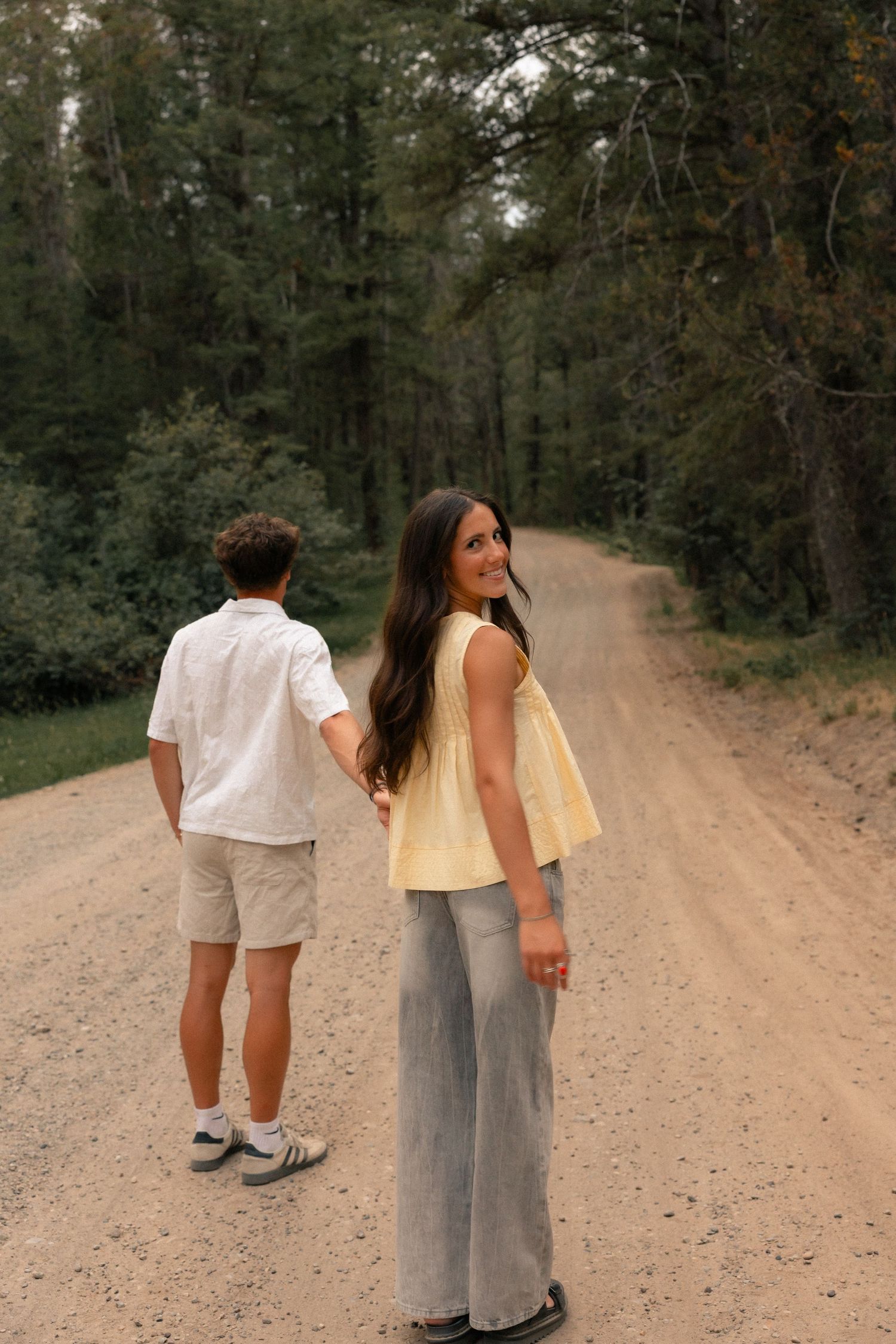 Couple holding hands on a dirt forest road, woman smiling back at camera.