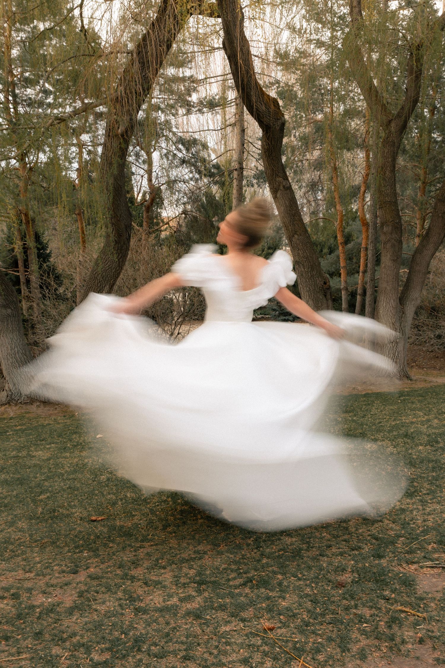 Bride in white gown spinning in forest, motion blur capturing flowing dress movement among tall trees.