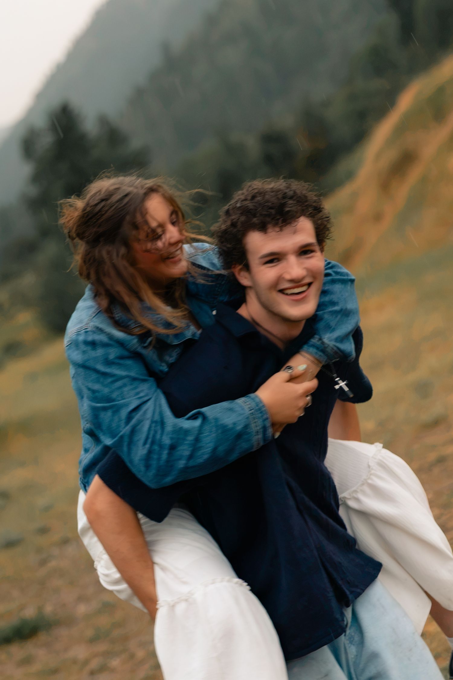A laughing young couple enjoys a piggyback ride in a misty mountain meadow, both dressed casually.
