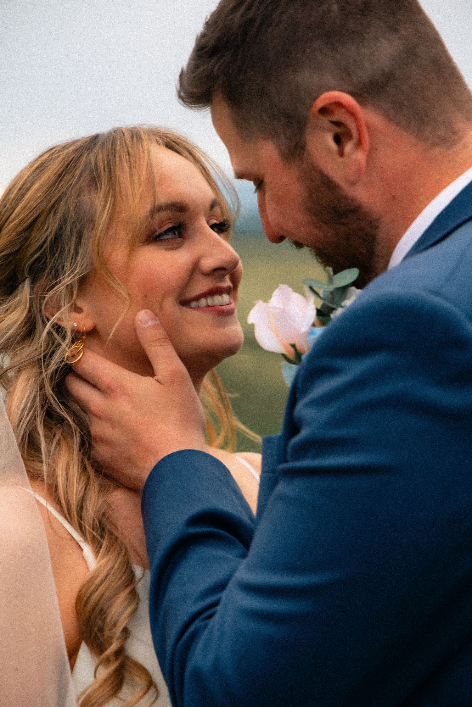 A groom in a blue suit tenderly holds his smiling bride's face during their romantic wedding portrait outdoors.