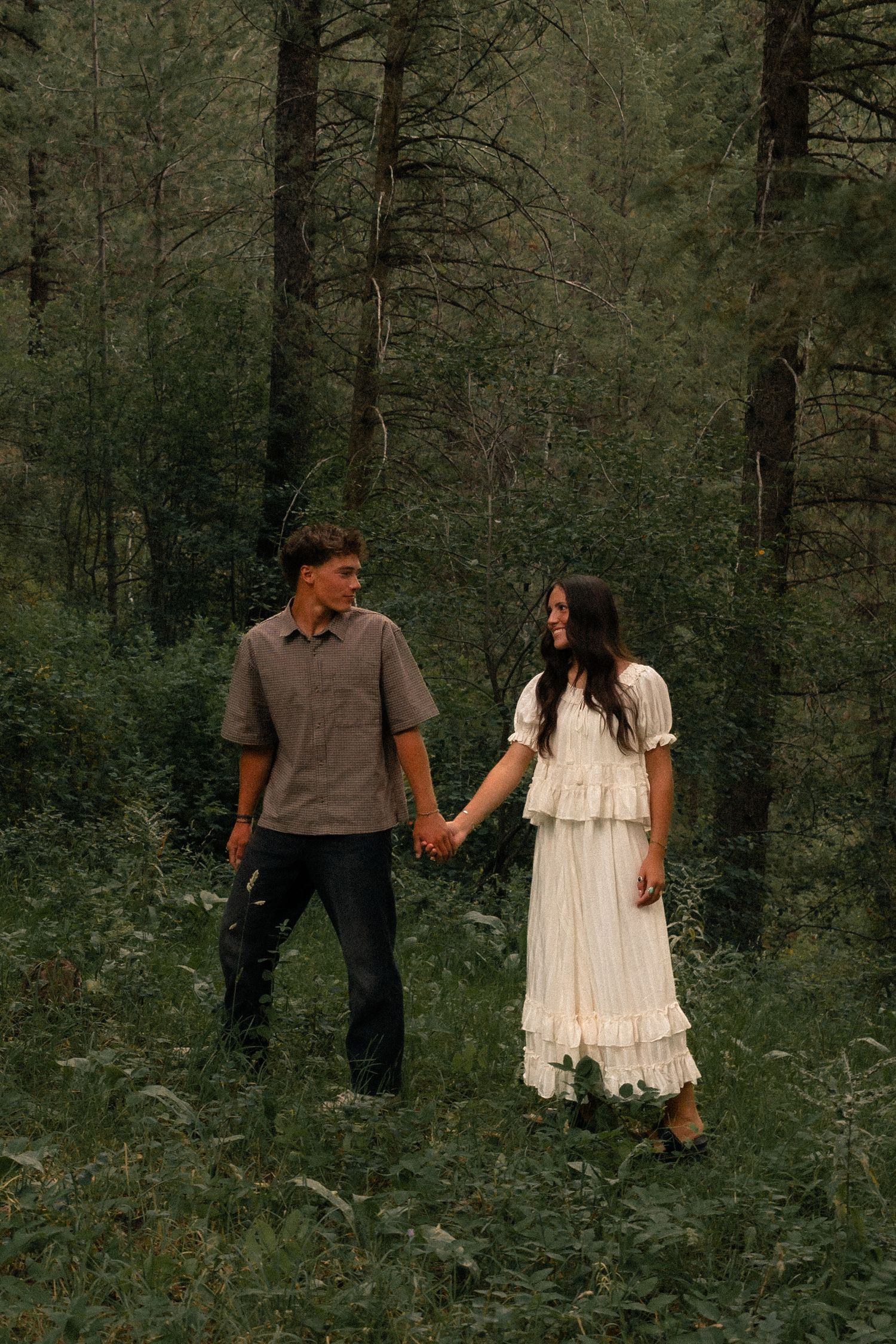 A couple holds hands while walking through a lush green forest, the woman in a white dress and man in a gray shirt.