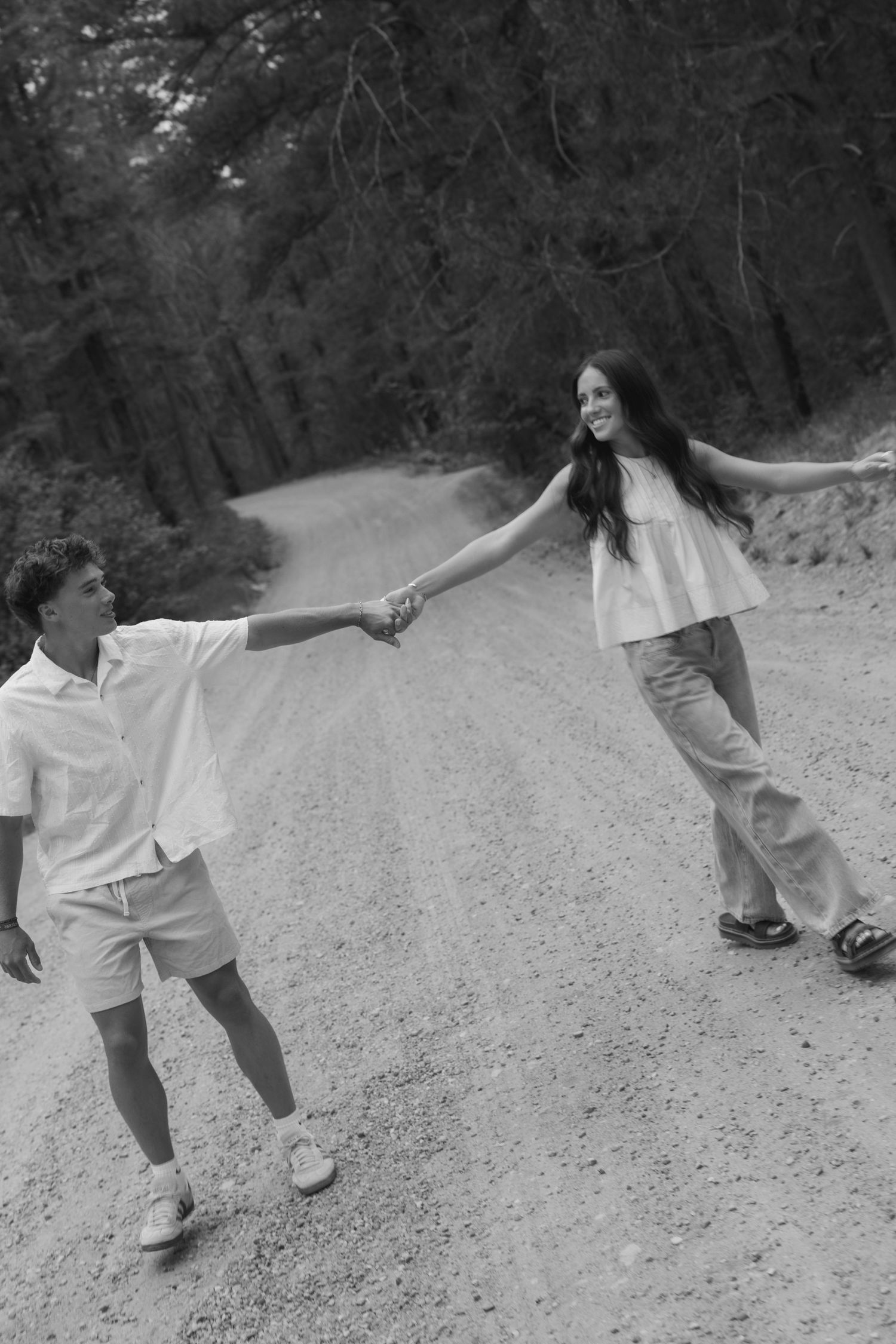 Black and white photo of a couple holding hands on a forest dirt road, smiling and spinning together.