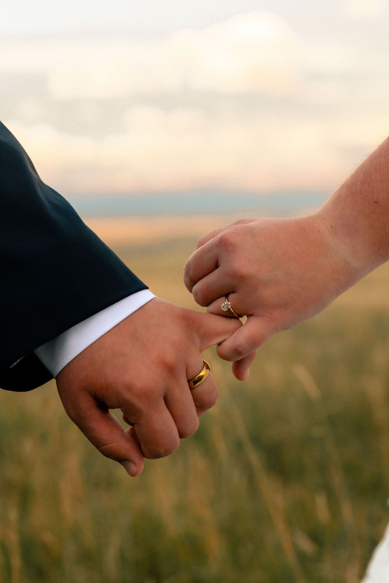 Close-up of newlywed couple holding hands showing gold wedding bands and diamond engagement ring outdoors.