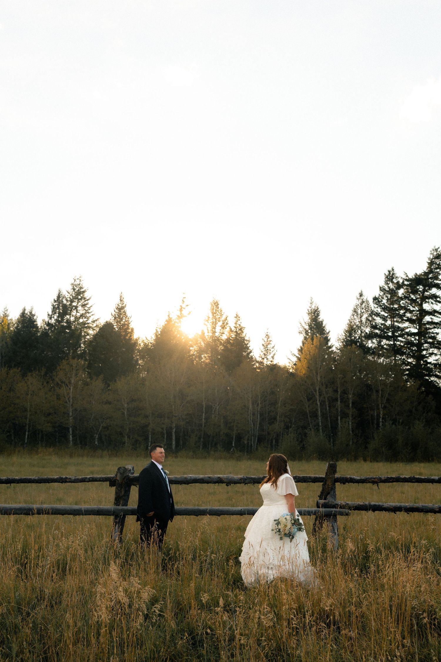 Bride and groom stand by a rustic wooden fence in a golden sunset field surrounded by tall evergreen trees.