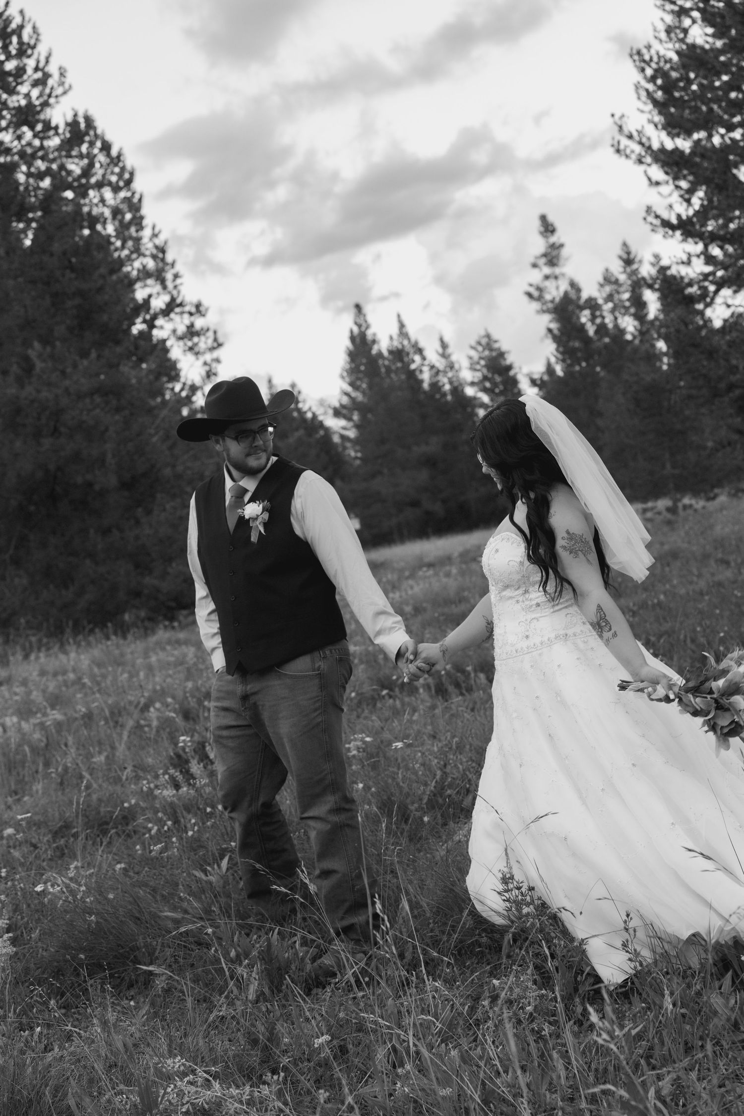 Black and white photo of a bride and groom holding hands walking through a field, groom wearing cowboy hat.
