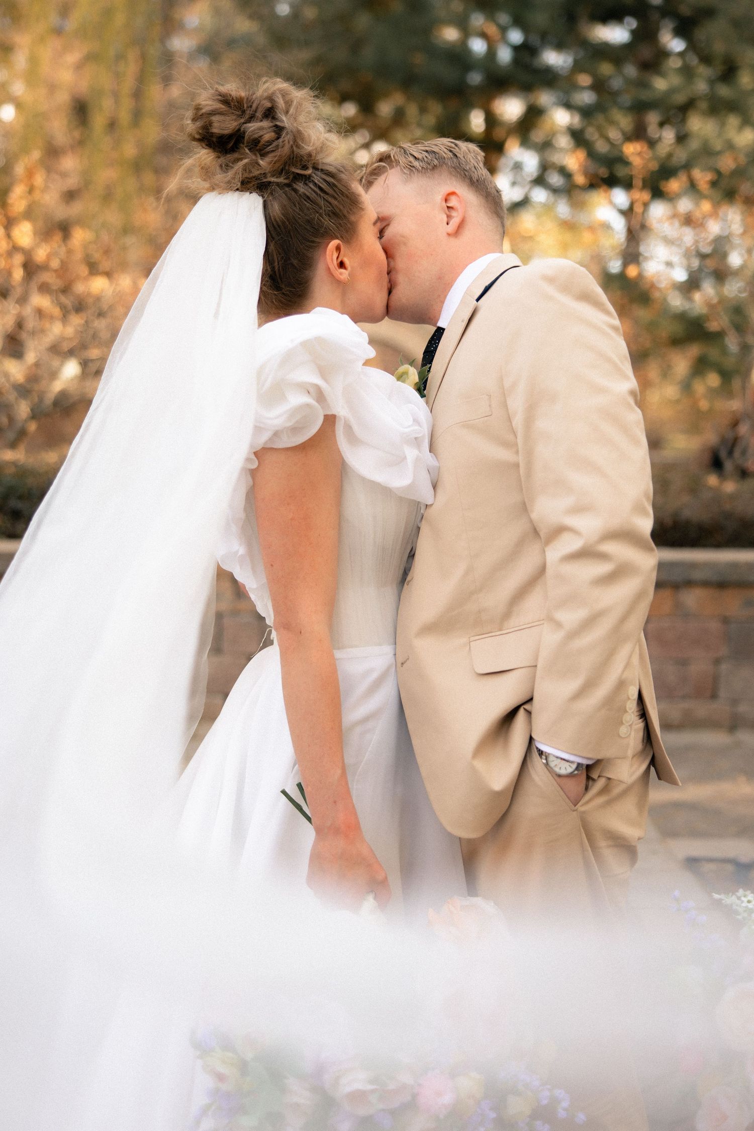 A bride in a white gown and veil shares a romantic kiss with a groom in a tan suit outdoors.