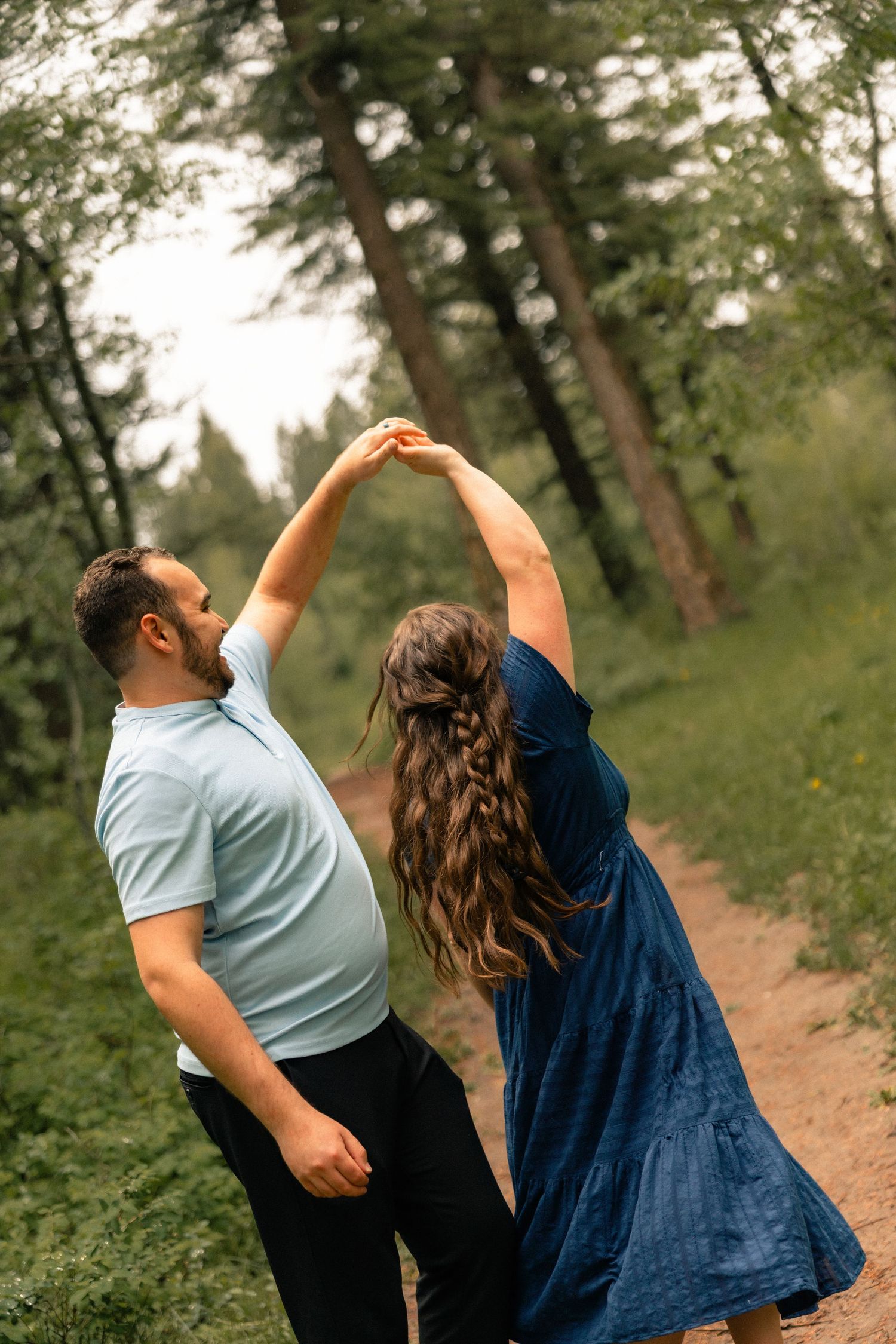 Couple dancing on a forest path, woman in blue dress spinning with arms raised, surrounded by tall trees.