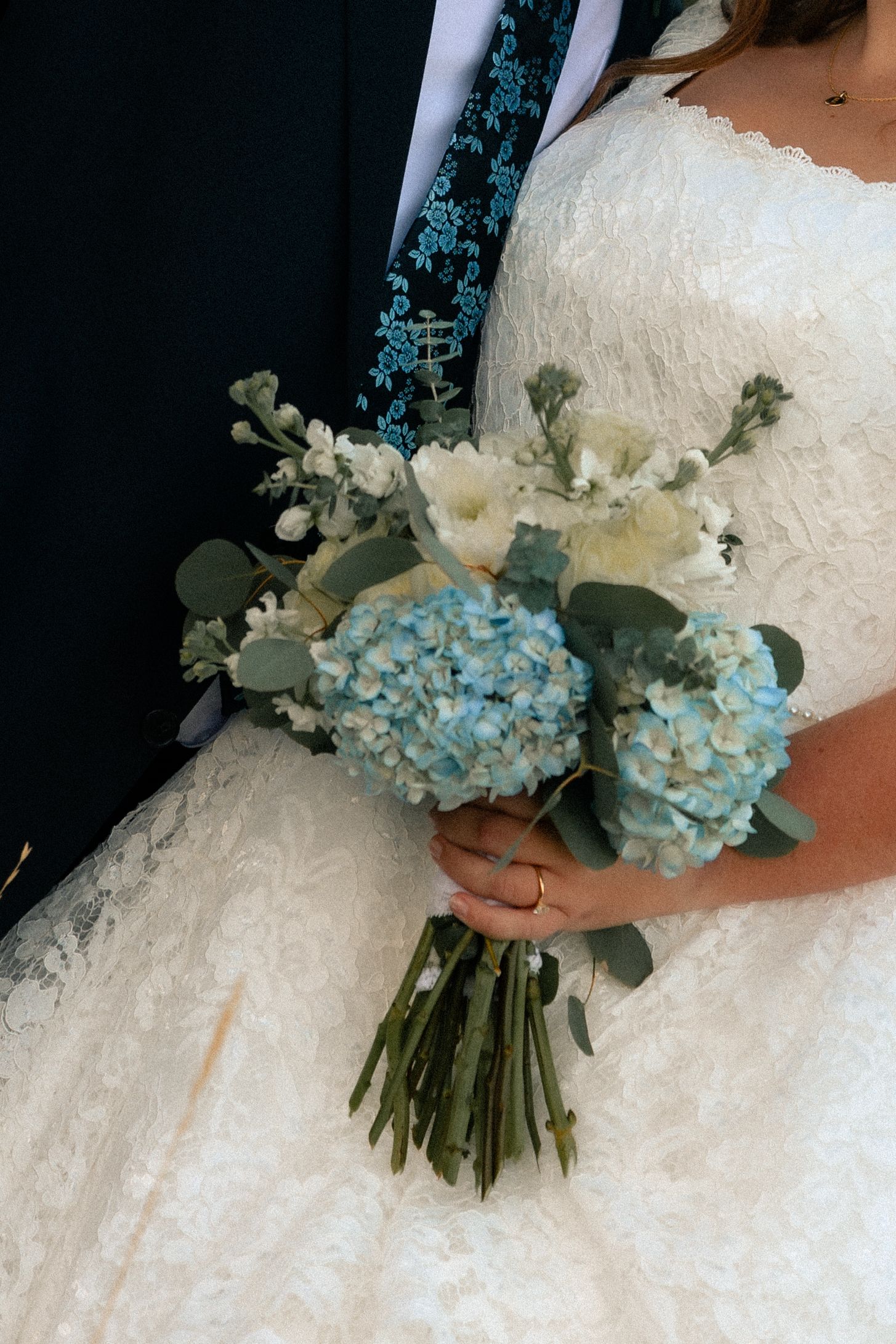 Bride in white lace gown holding a bouquet of blue hydrangeas, white flowers, and eucalyptus greenery.