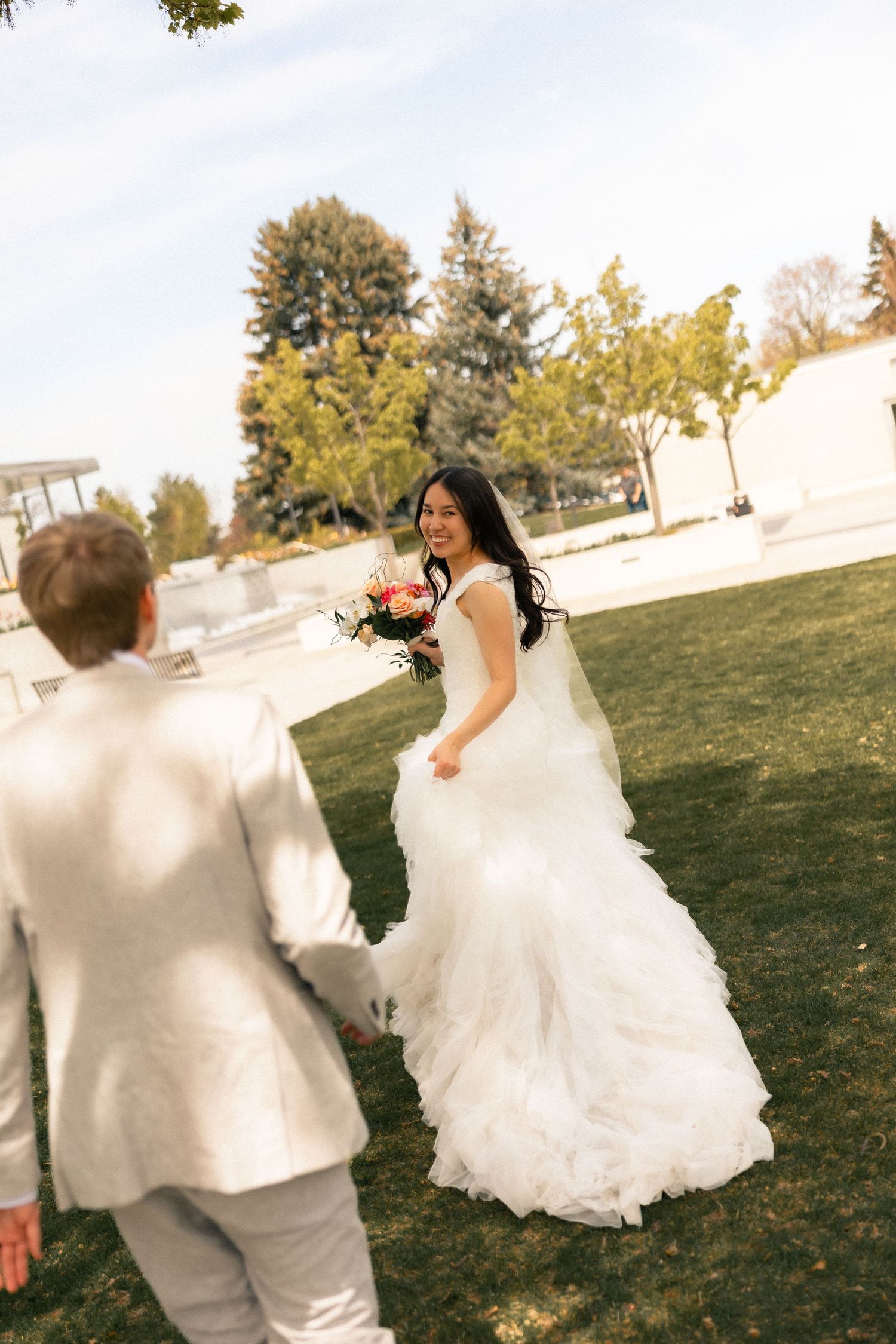 Bride in white ruffled gown holding bouquet smiles at groom in ivory suit on green lawn with trees.