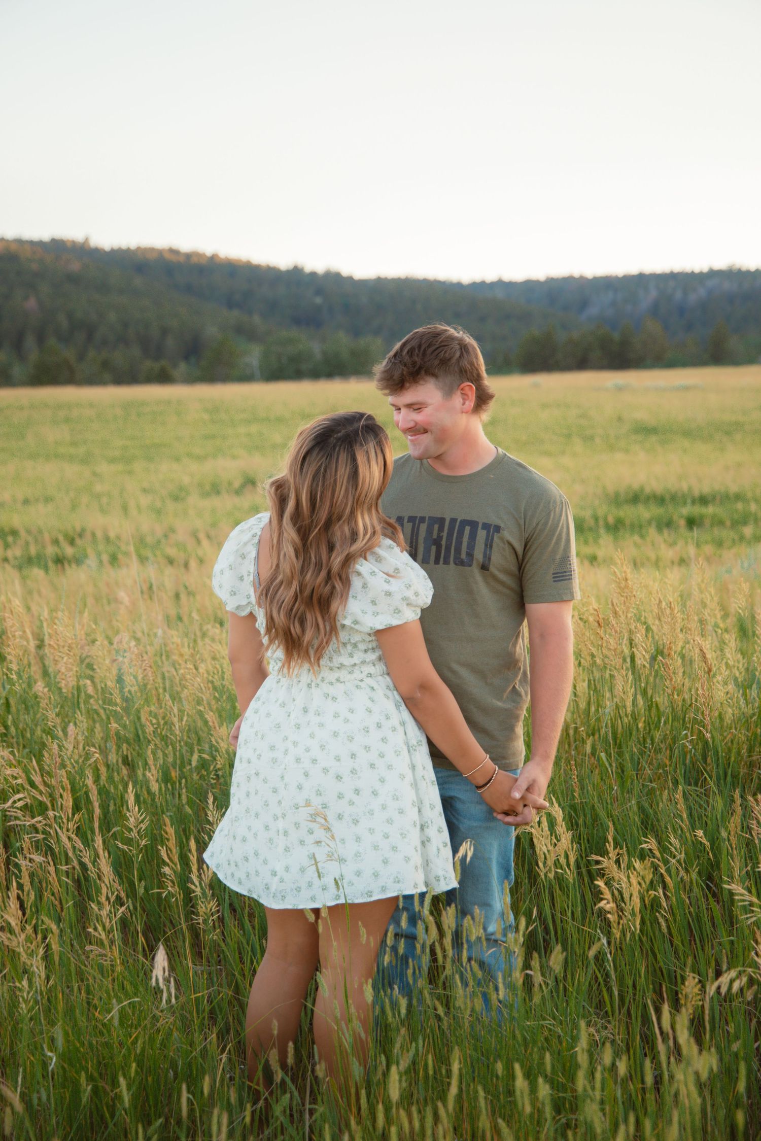 A couple shares a playful moment in a summer field with rolling hills in the background.