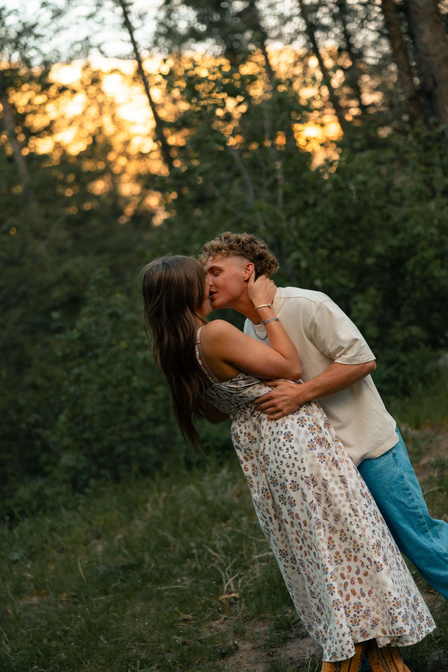 Romantic couple sharing a kiss in a forest clearing with golden sunset light filtering through the trees behind them.