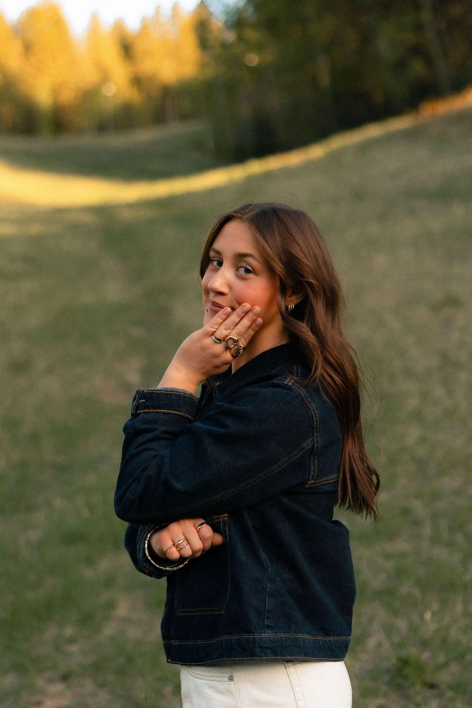 Young woman in denim jacket poses outdoors at golden hour with trees and green hillside in background.