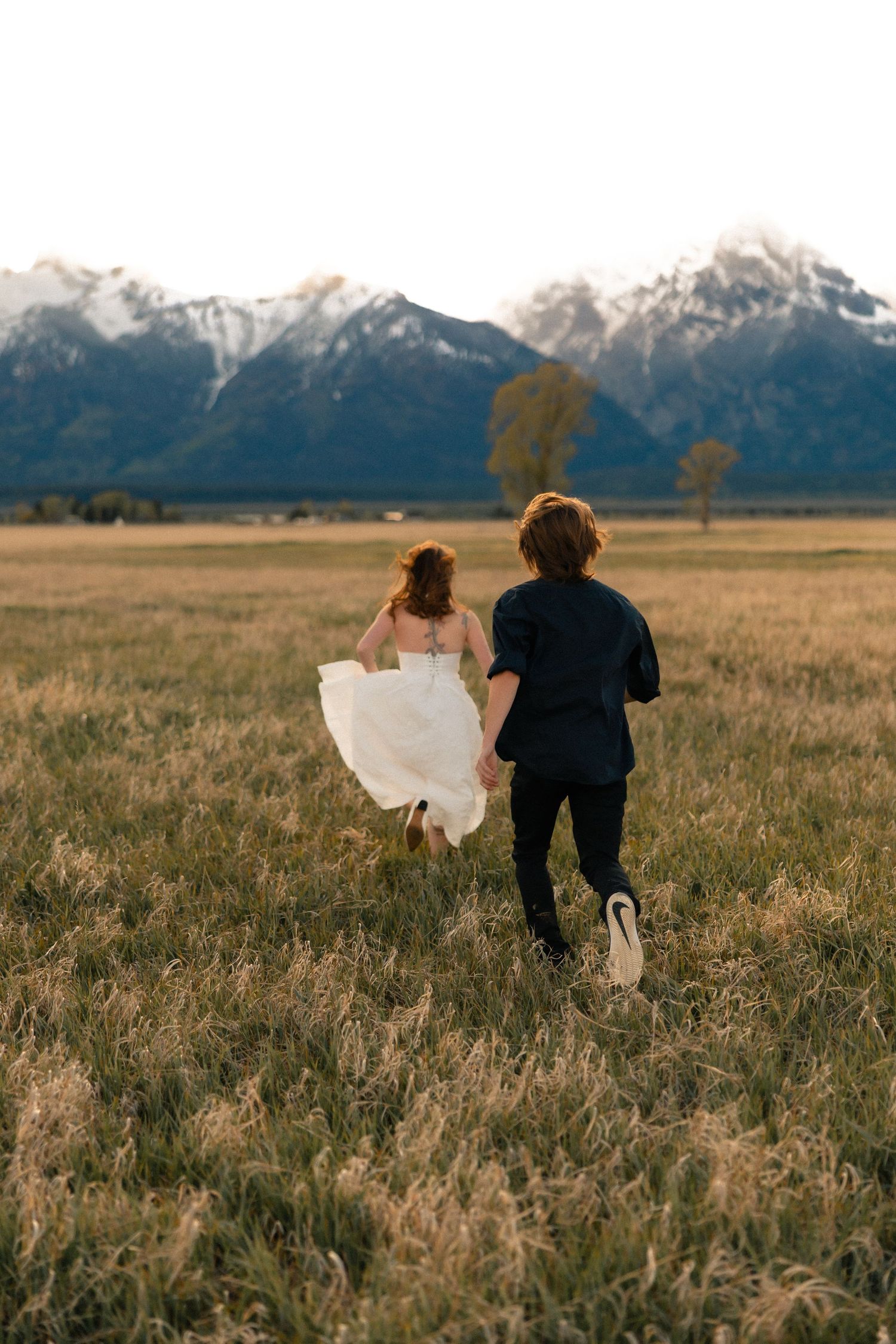 A bride and groom run hand-in-hand through a golden meadow with snow-capped Grand Teton mountains in the background.