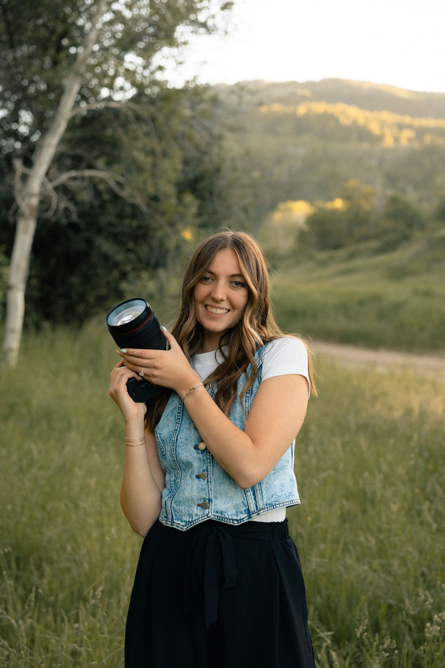 Smiling young woman holding a camera lens outdoors in a green meadow with trees and hills at golden hour.