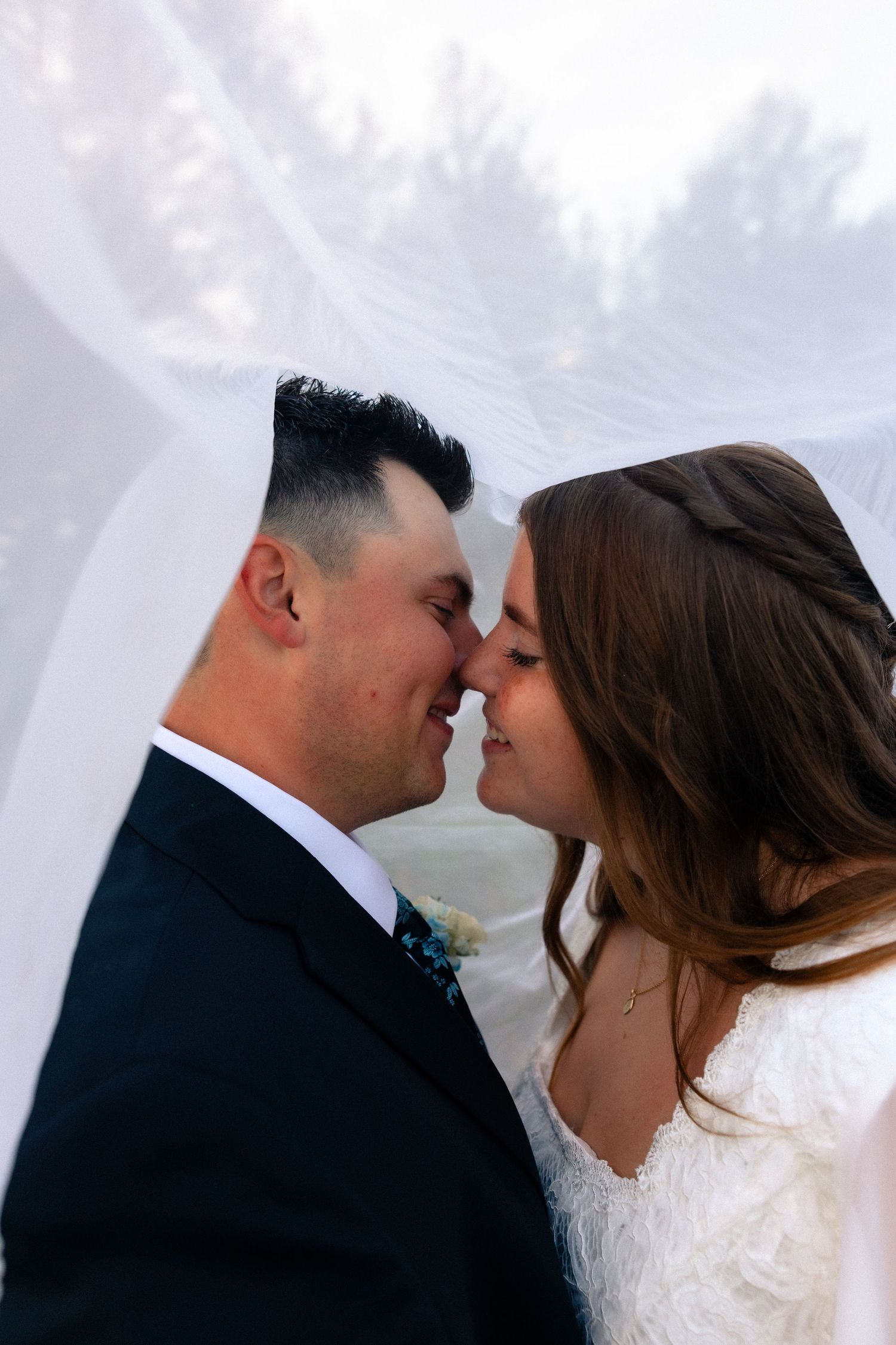 Bride and groom sharing a romantic almost-kiss under a flowing white veil on their wedding day.