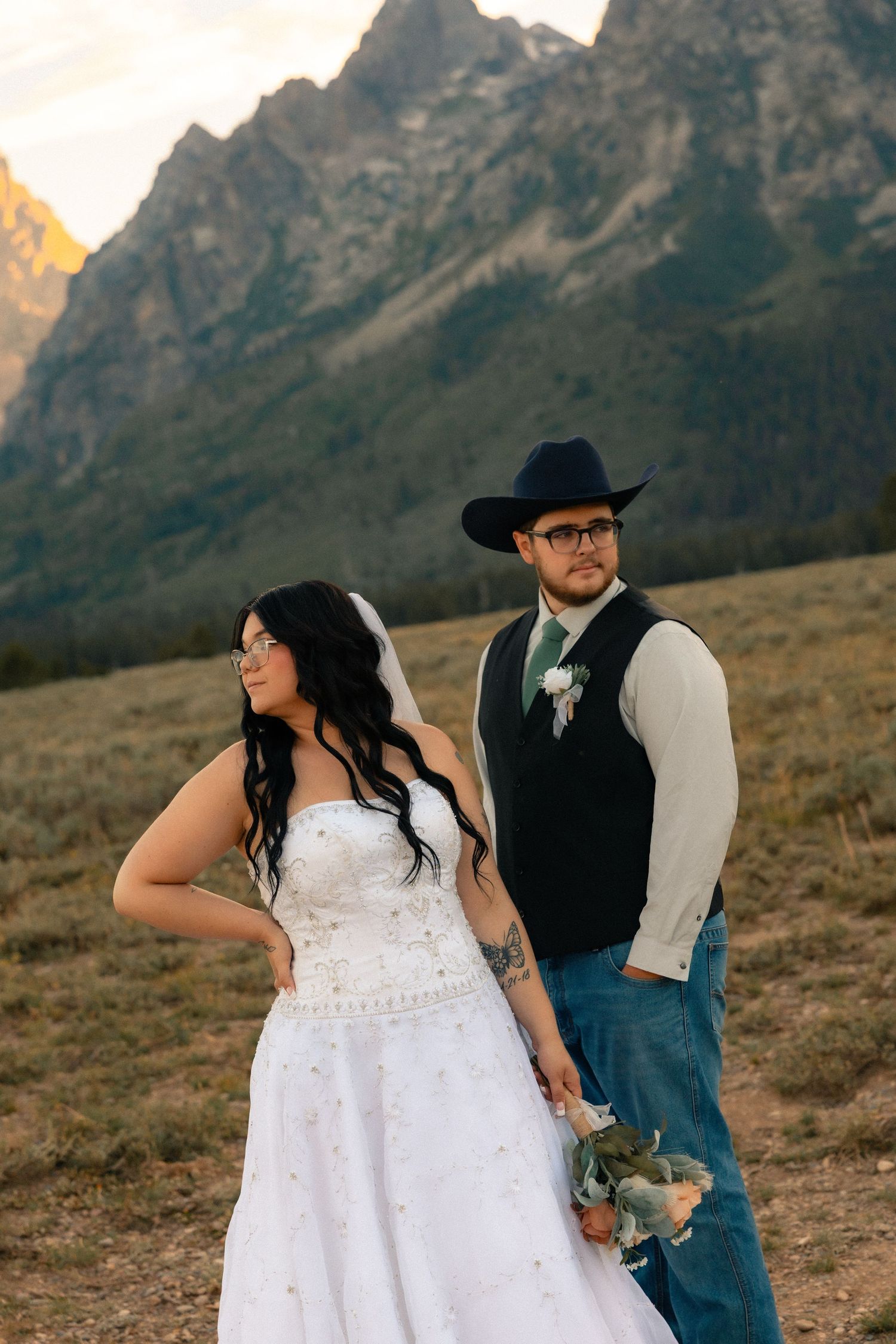 Bride in white gown and groom in cowboy hat pose before Grand Teton mountains at sunset wedding.