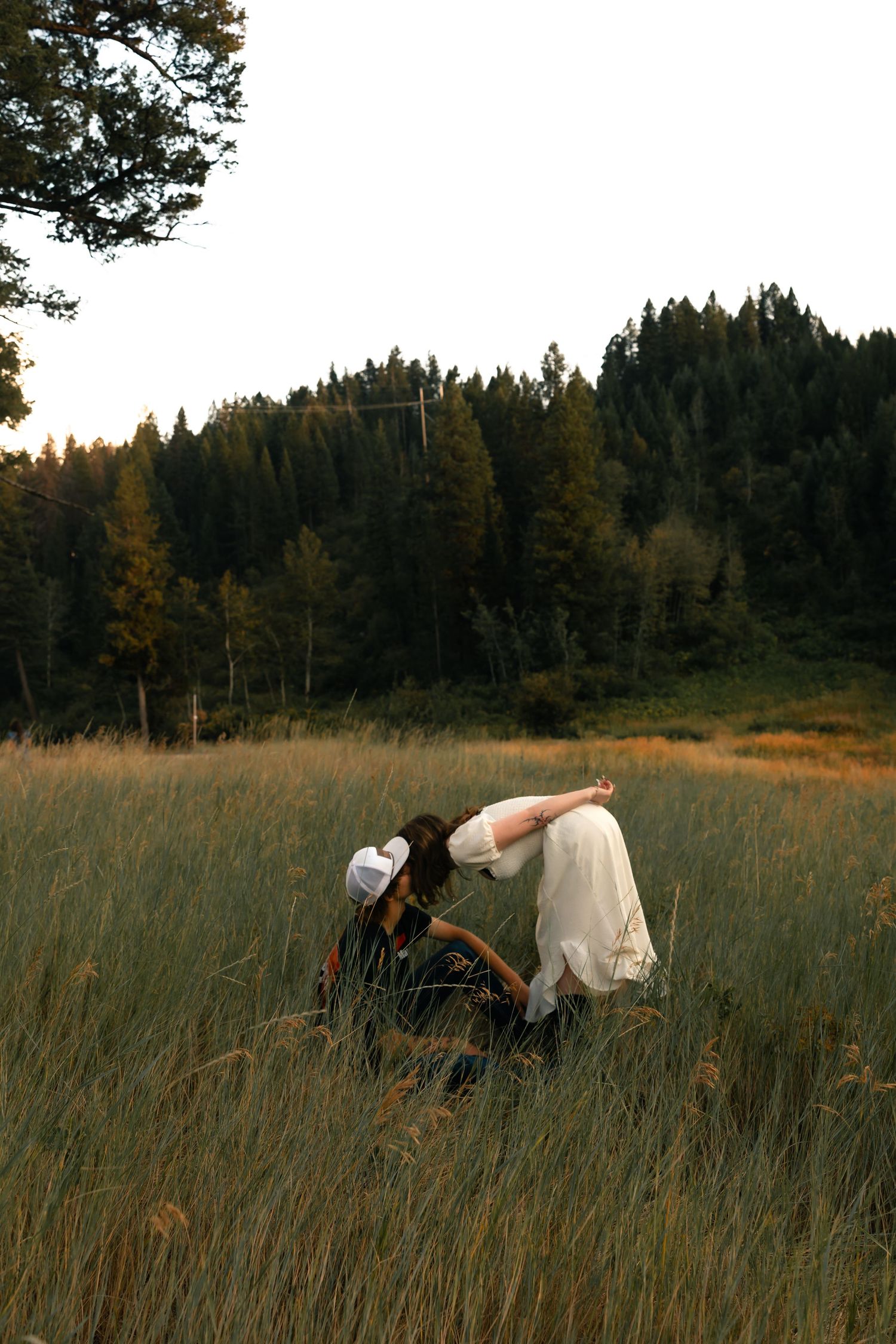 Two people in white and dark clothing embrace in a tall grass meadow surrounded by evergreen forest at golden hour.