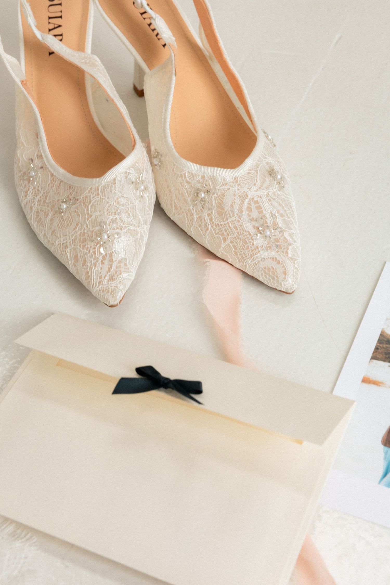 Elegant ivory lace bridal heels with pearl embellishments beside a cream envelope with black ribbon bow.