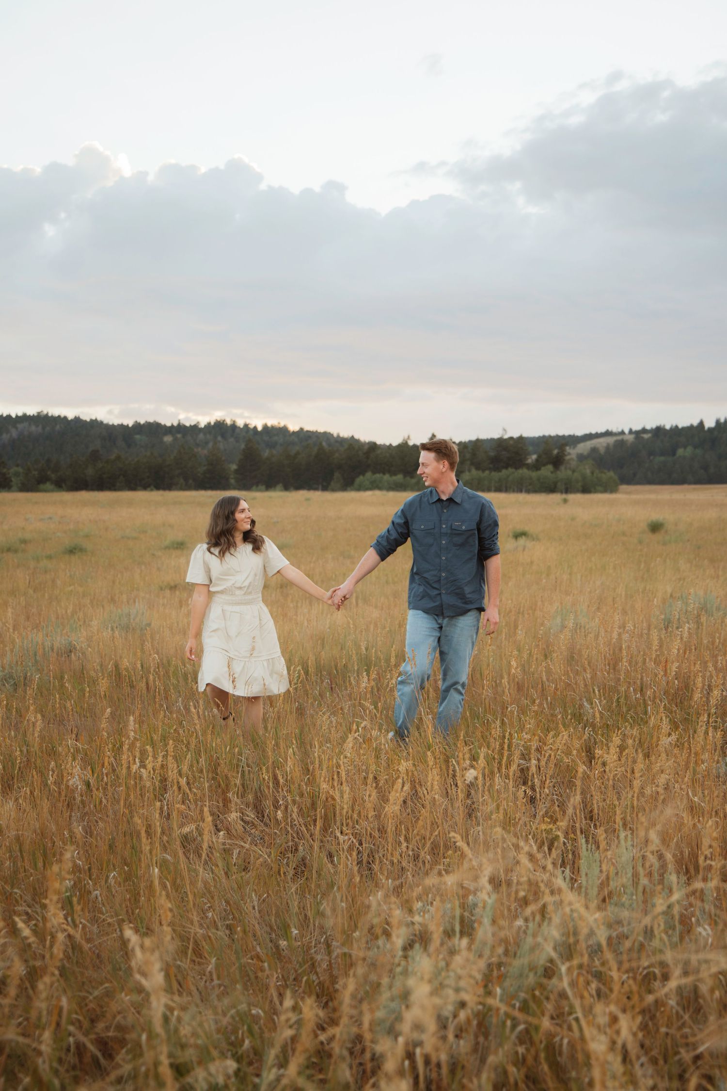 Couple holding hands while walking through a golden wheat field at dusk.