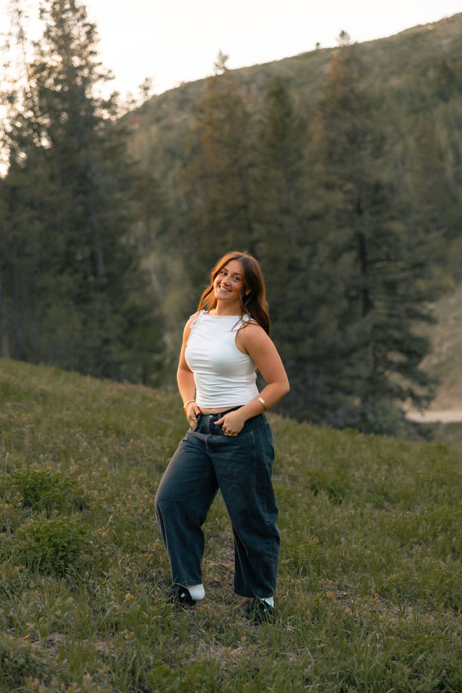A young woman in a white tank top and dark jeans poses on a grassy hillside surrounded by pine trees.