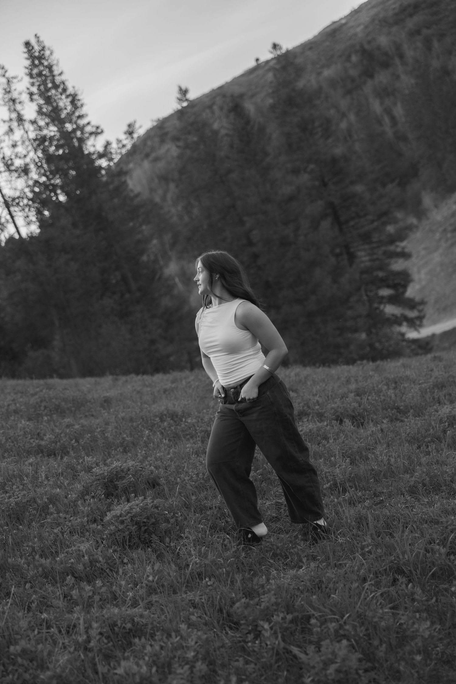 Black and white photo of a young woman in a white tank top and dark jeans standing on a grassy hillside.