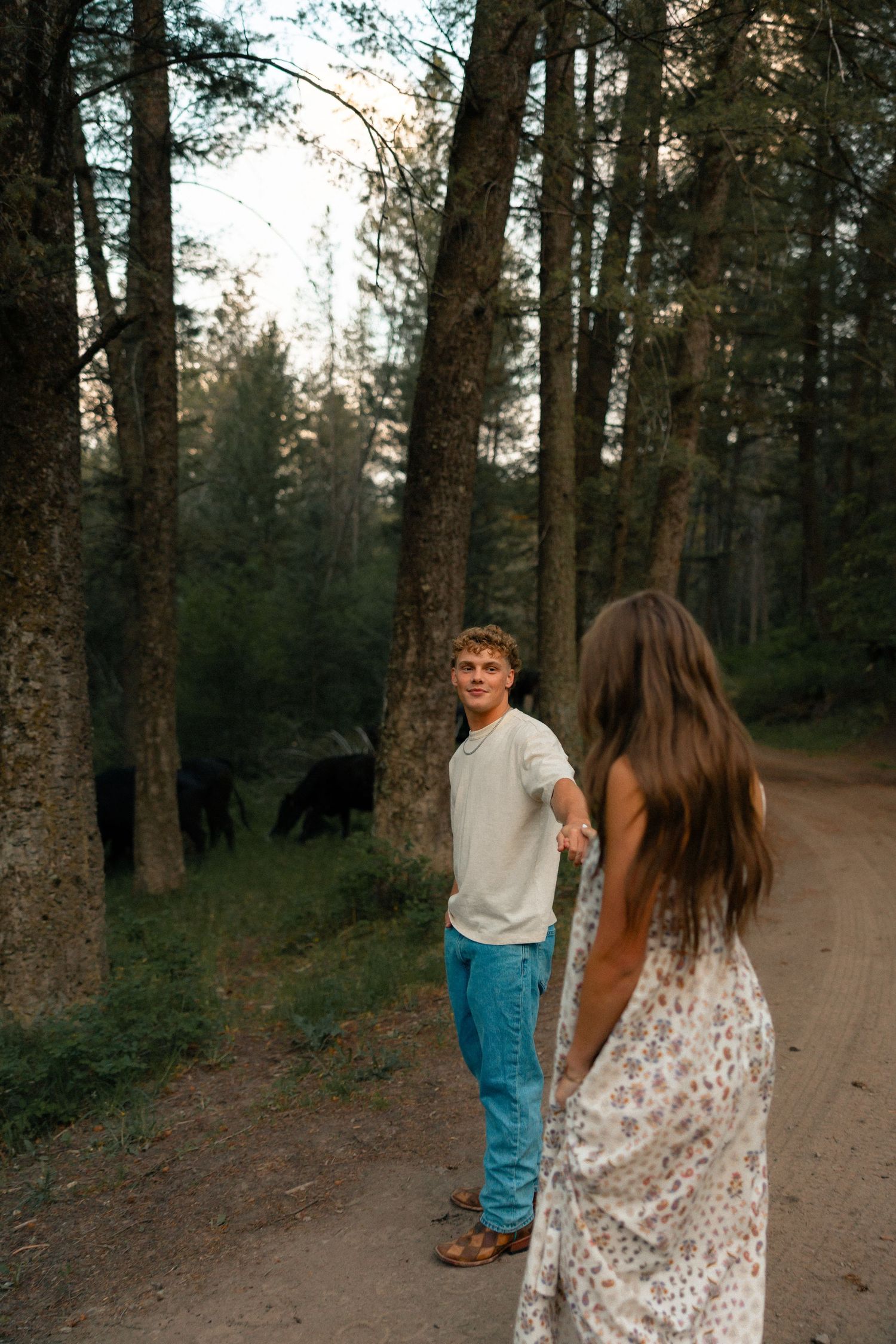 A young couple holds hands on a forest path at dusk, with a black bear visible among the trees behind them.