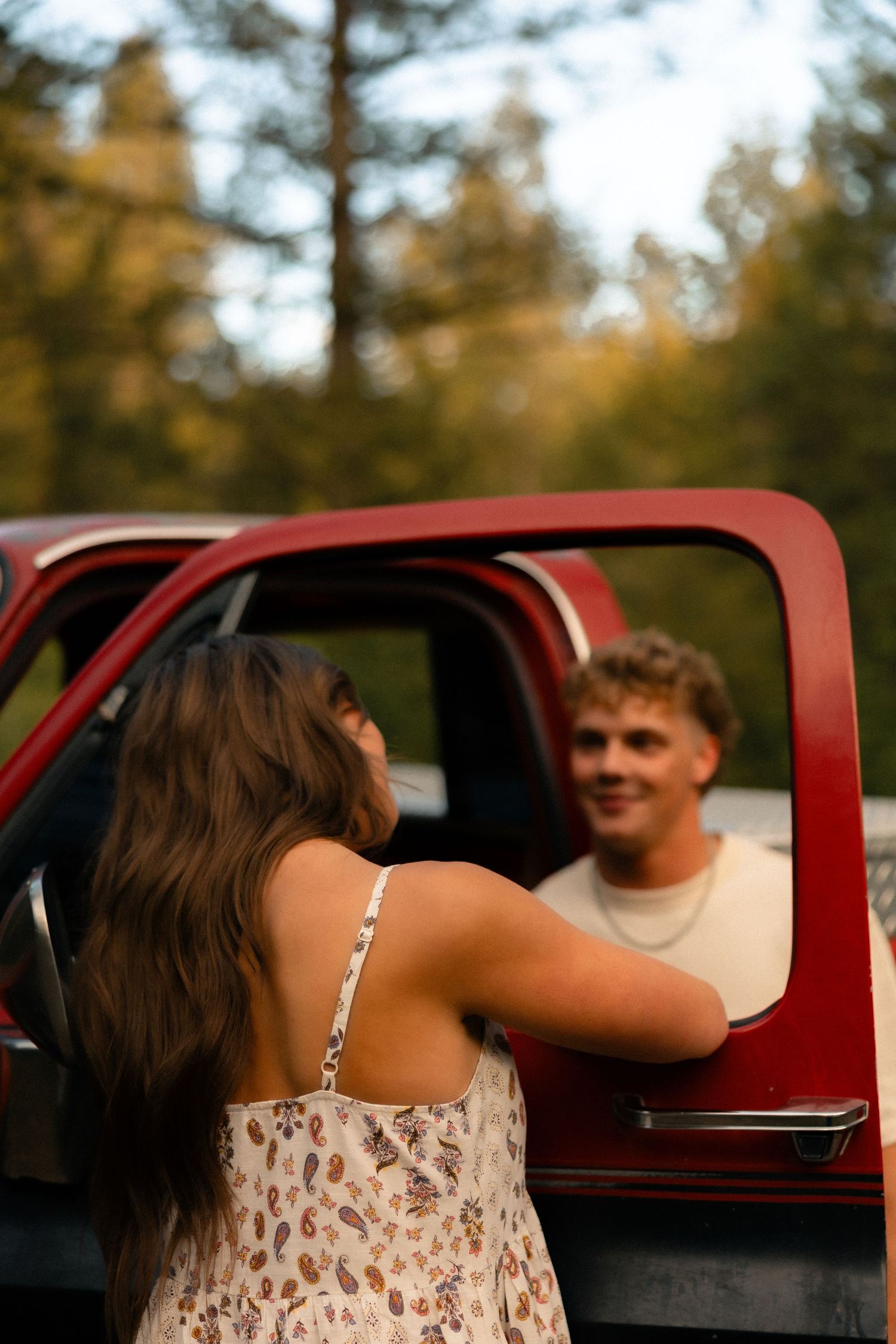 A young couple shares a smile near a red truck surrounded by autumn trees.