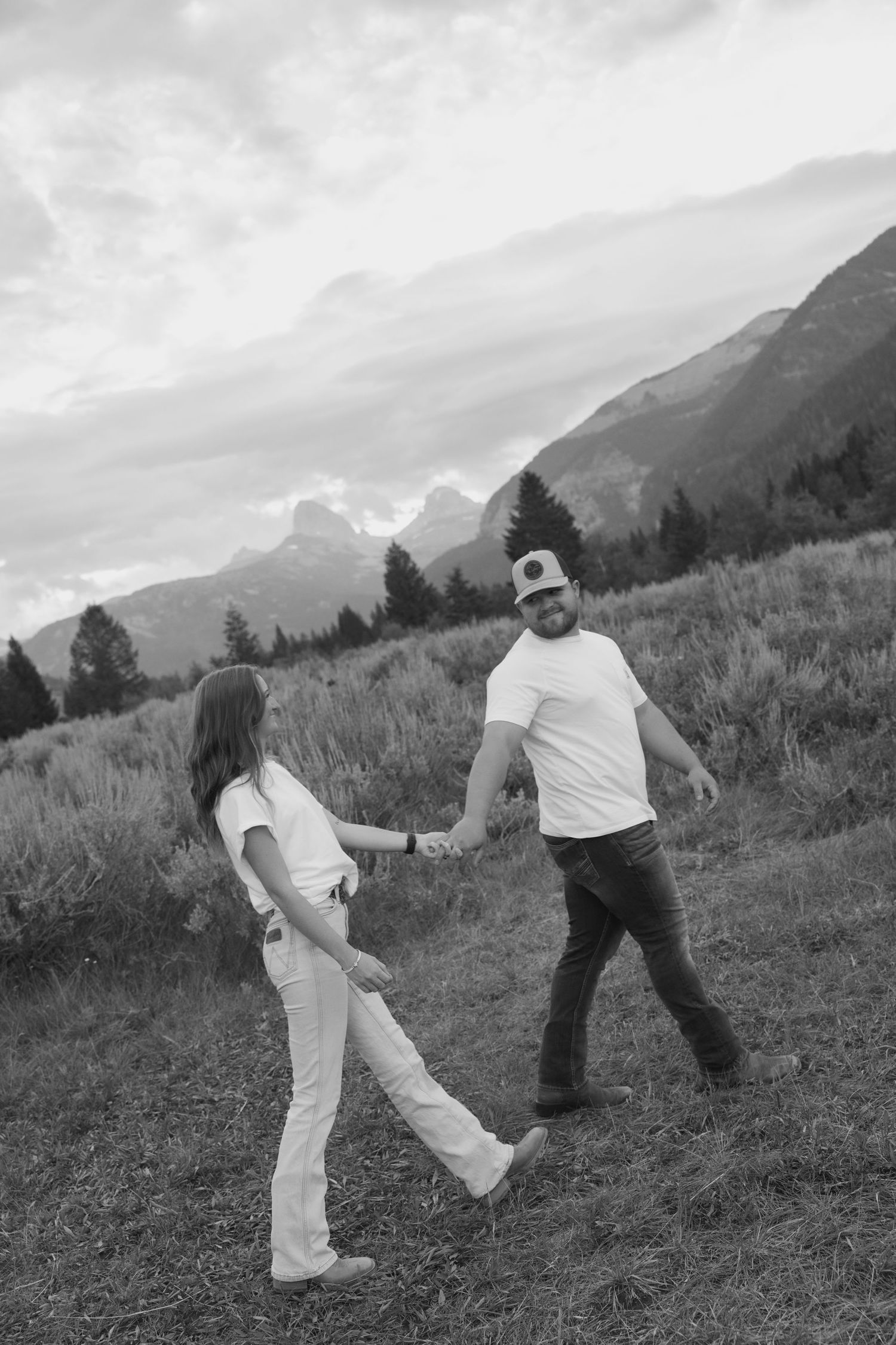 Black and white photo of a couple holding hands while walking through a mountain meadow with scenic peaks behind them.