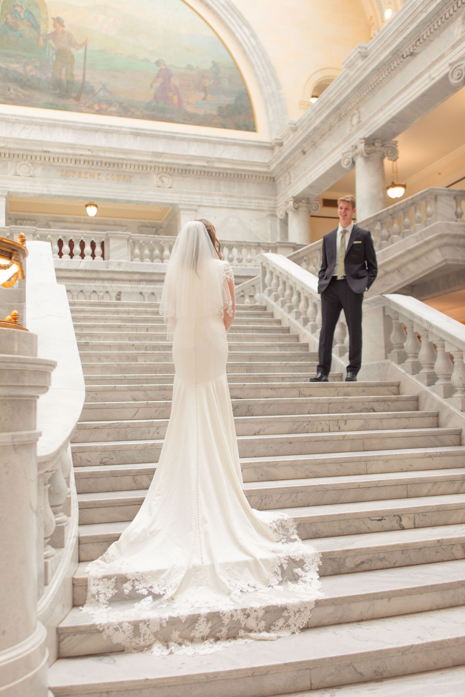 Bride in white gown with lace veil ascending marble staircase toward groom in dark suit at elegant wedding venue.
