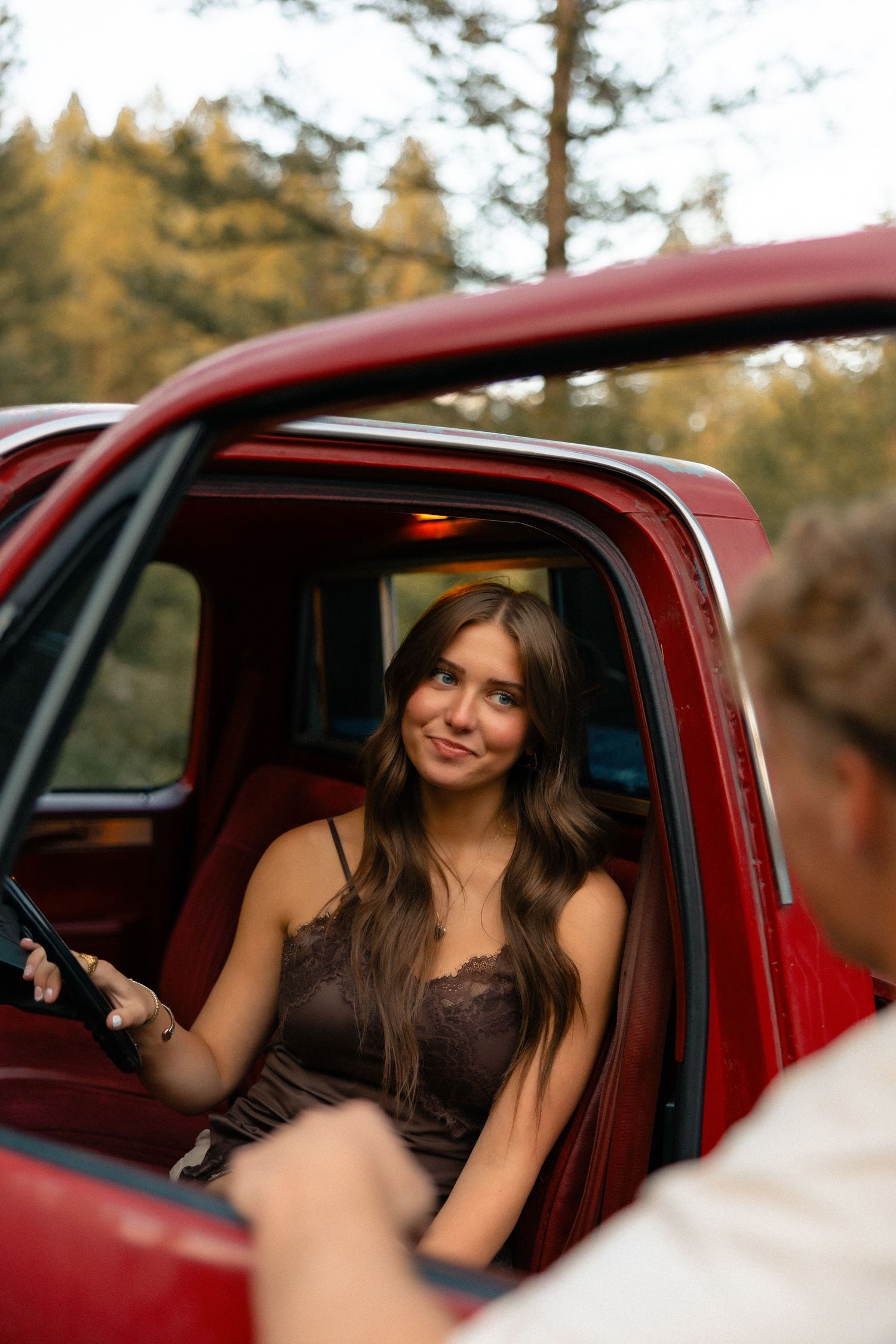 Young woman in brown lace dress smiling from red vintage truck cab in forest setting.