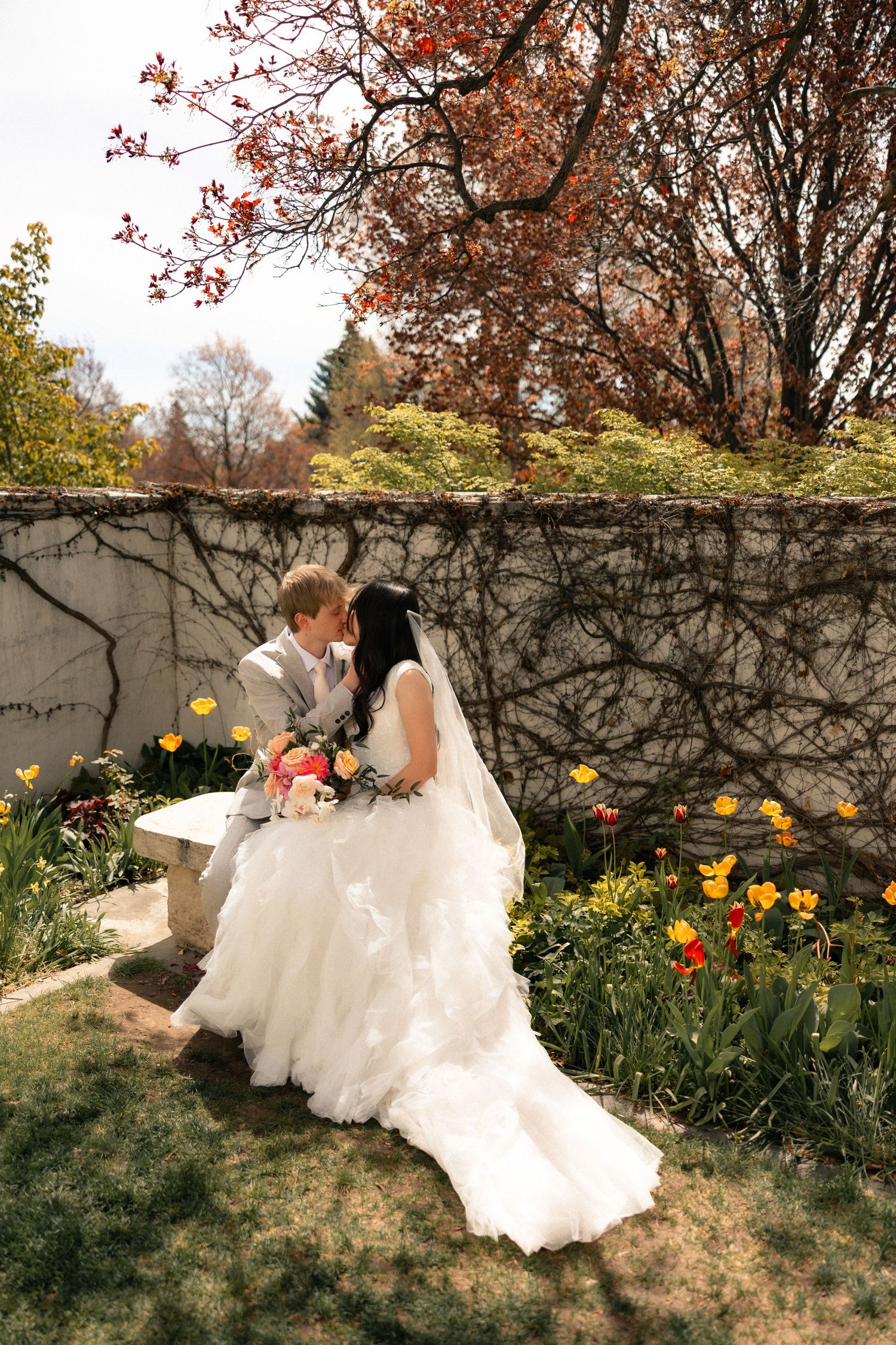 Bride and groom share a kiss on a garden bench surrounded by colorful tulips and autumn foliage on their wedding day.