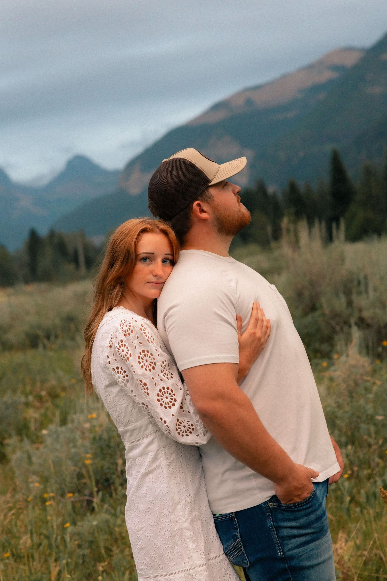 Couple embracing in a mountain meadow, woman in white lace dress leaning on man in white tee and cap.