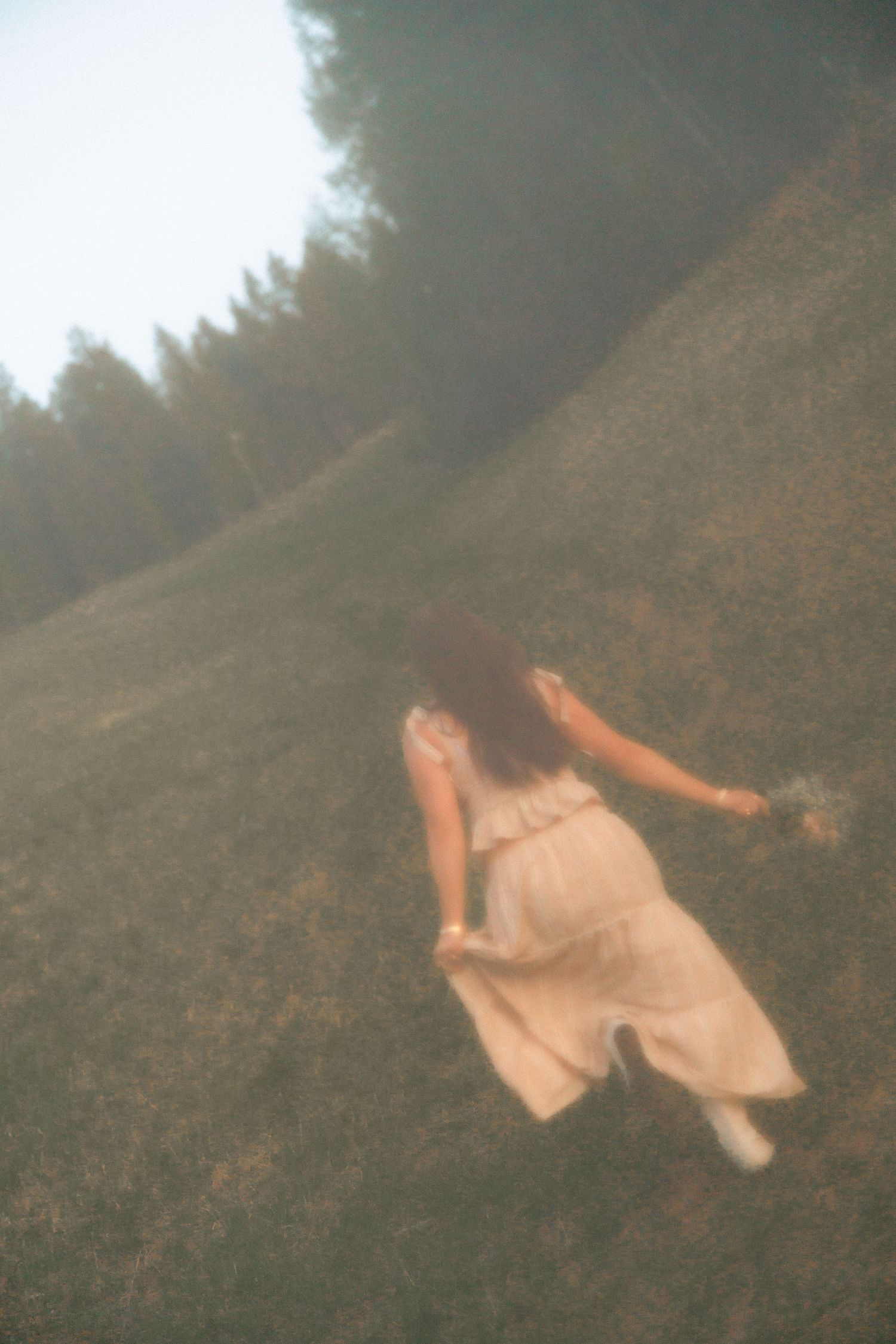 Woman in white dress running up a grassy hillside with trees, dreamy blurred film photography style.