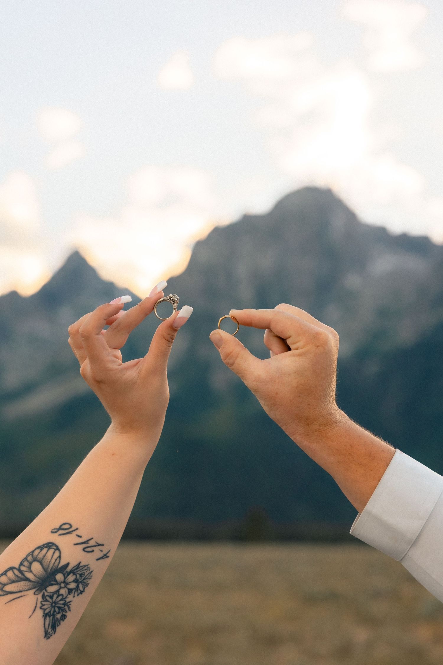 Two hands holding wedding rings with mountain backdrop, one with floral tattoo and white nails, other in white dress shirt sleeve.