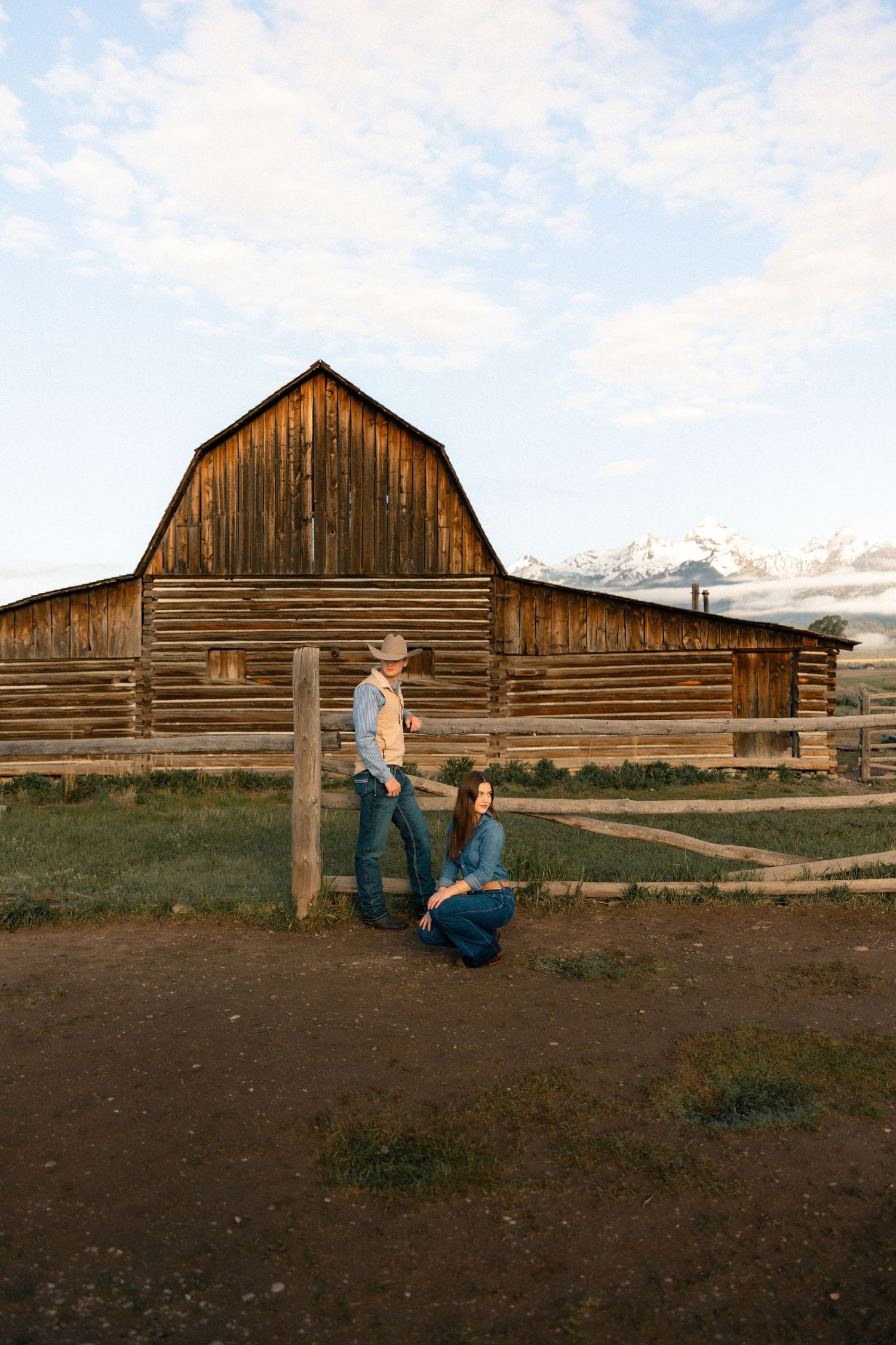 Couple posing by rustic wooden barn with Grand Teton mountains backdrop, man in cowboy hat standing, woman crouching.