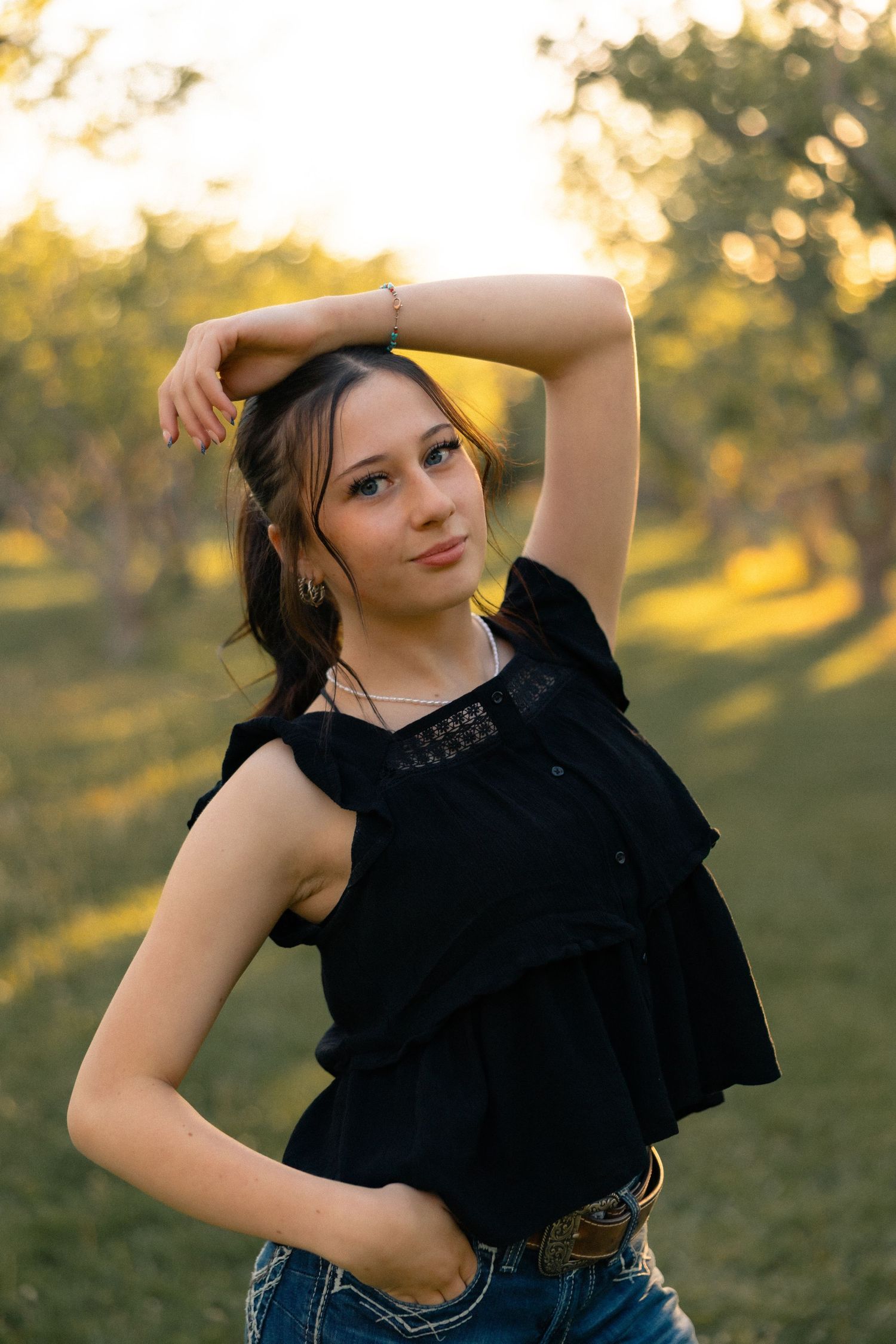 Young woman in black ruffled top and jeans poses outdoors at golden hour with hand raised above head.