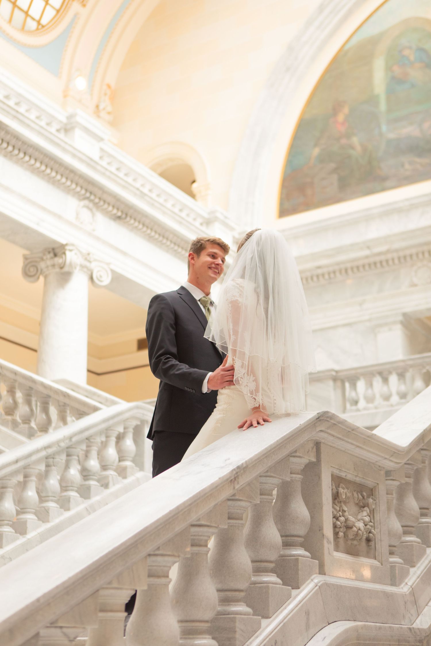 Smiling groom in dark suit stands beside bride in white veil on grand marble staircase at classic wedding venue.
