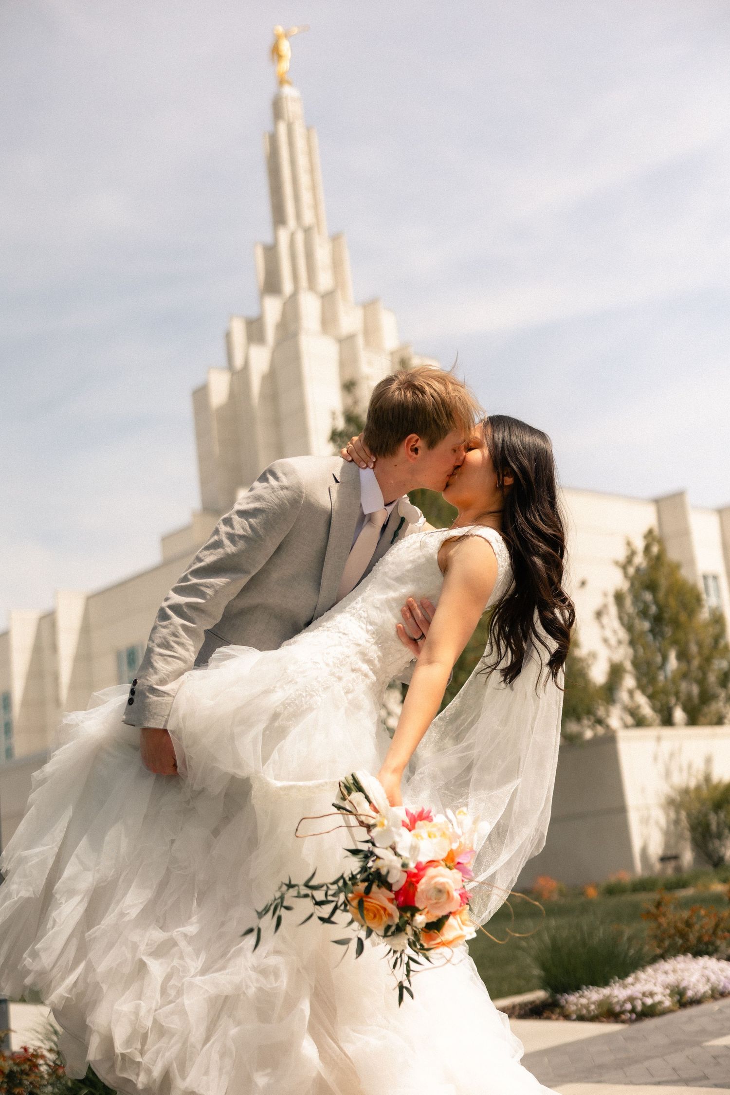 Bride and groom kissing in front of an LDS temple, bride holding a colorful bouquet in a white tulle gown.
