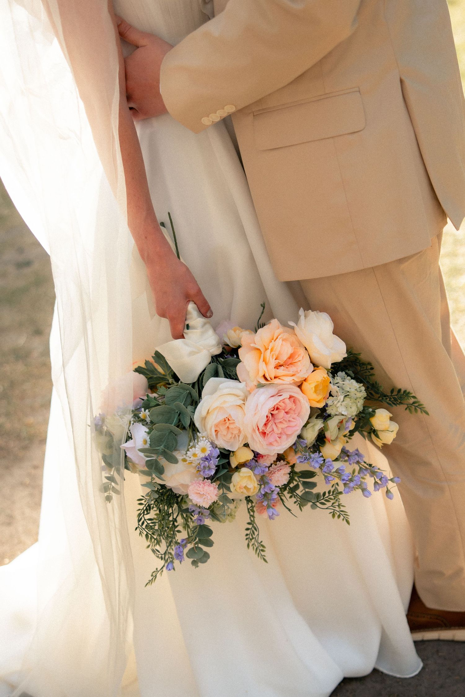 Bride holding a pastel wedding bouquet of peach roses, white flowers, and greenery while standing with groom in beige suit.