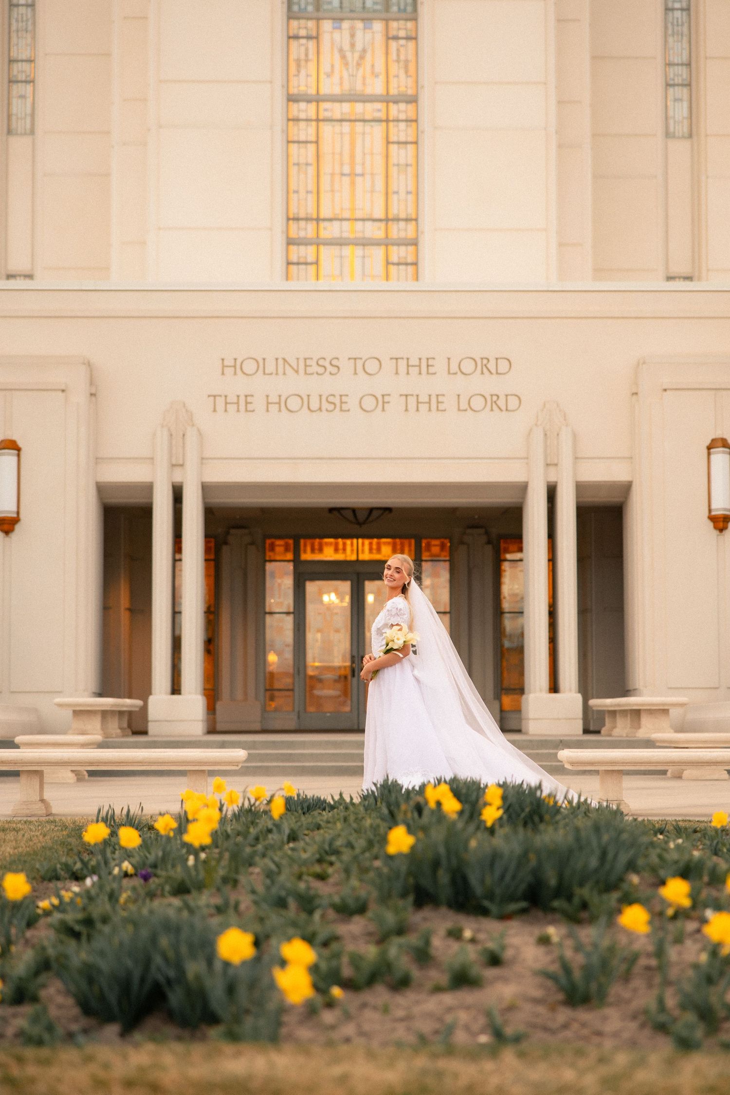 Bride in white gown and veil stands before LDS temple entrance with yellow daffodils blooming in foreground.