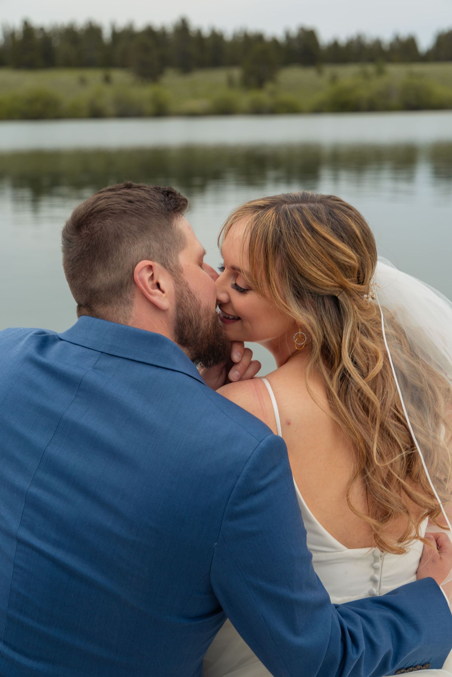 Bride and groom sharing a romantic kiss by a serene lake, with the groom in a blue suit and bride in a white gown.